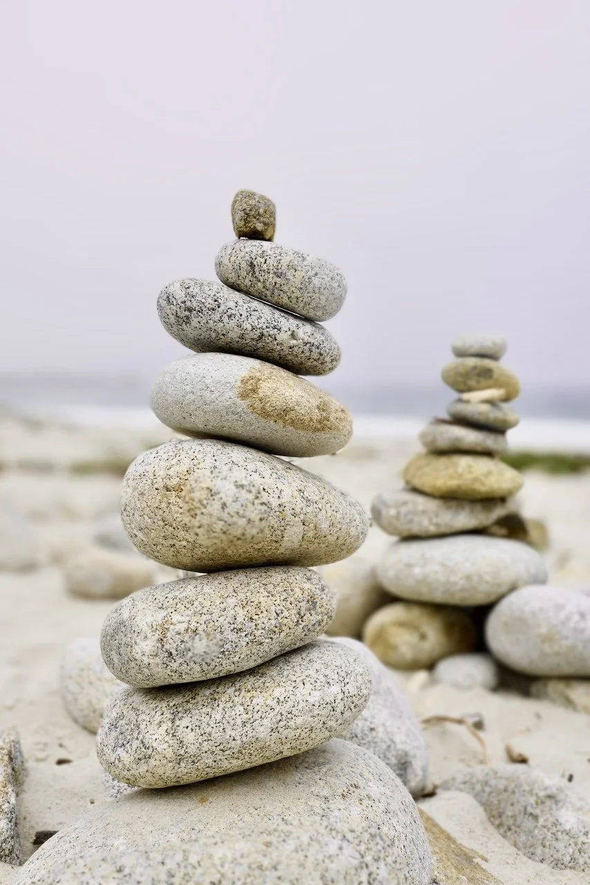 Two stacks of balanced beach stones on sandy ground with a blurred background of the beach and ocean.