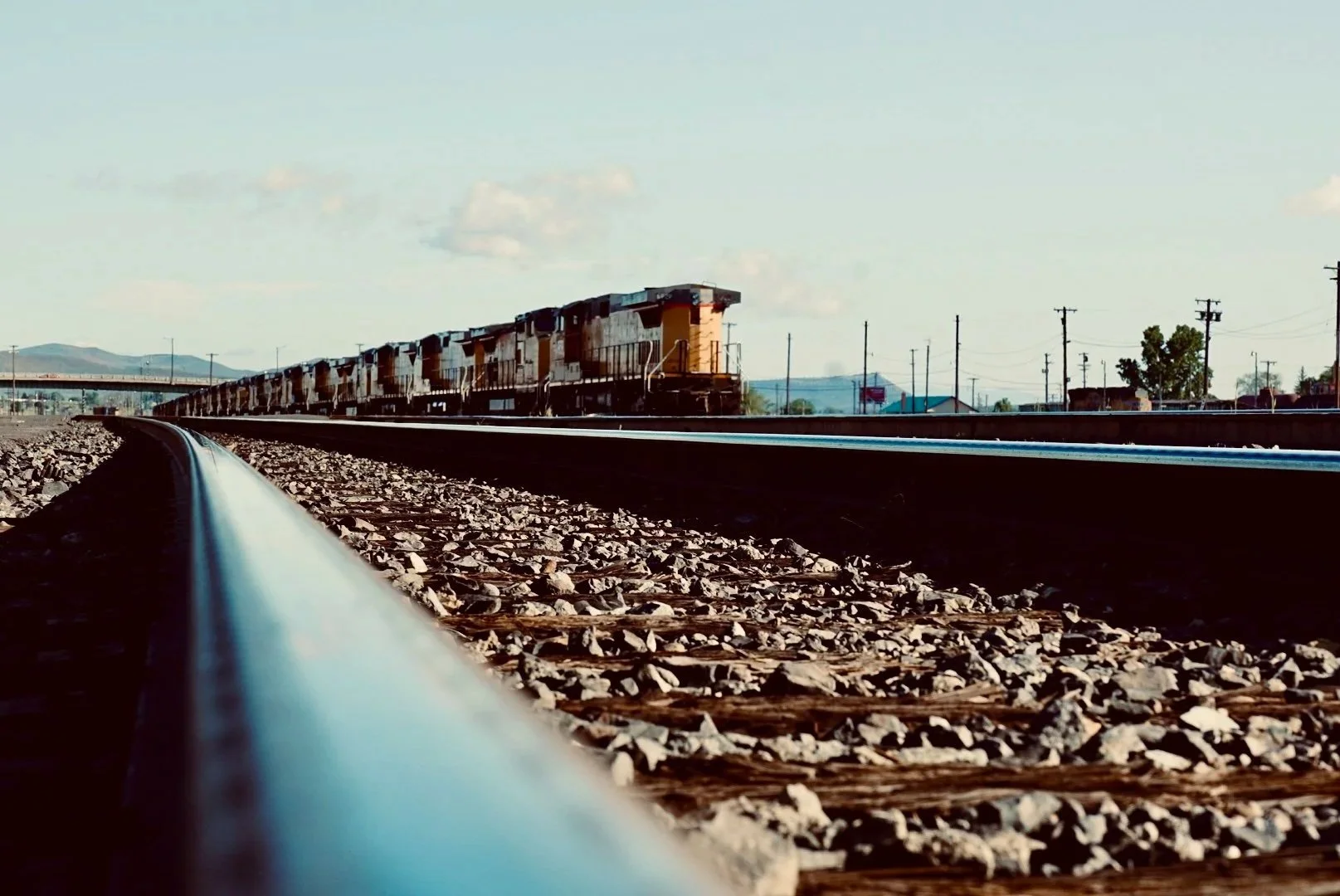 A low-angle view of a moving freight train on train tracks, with utility poles and a distant landscape in the background.