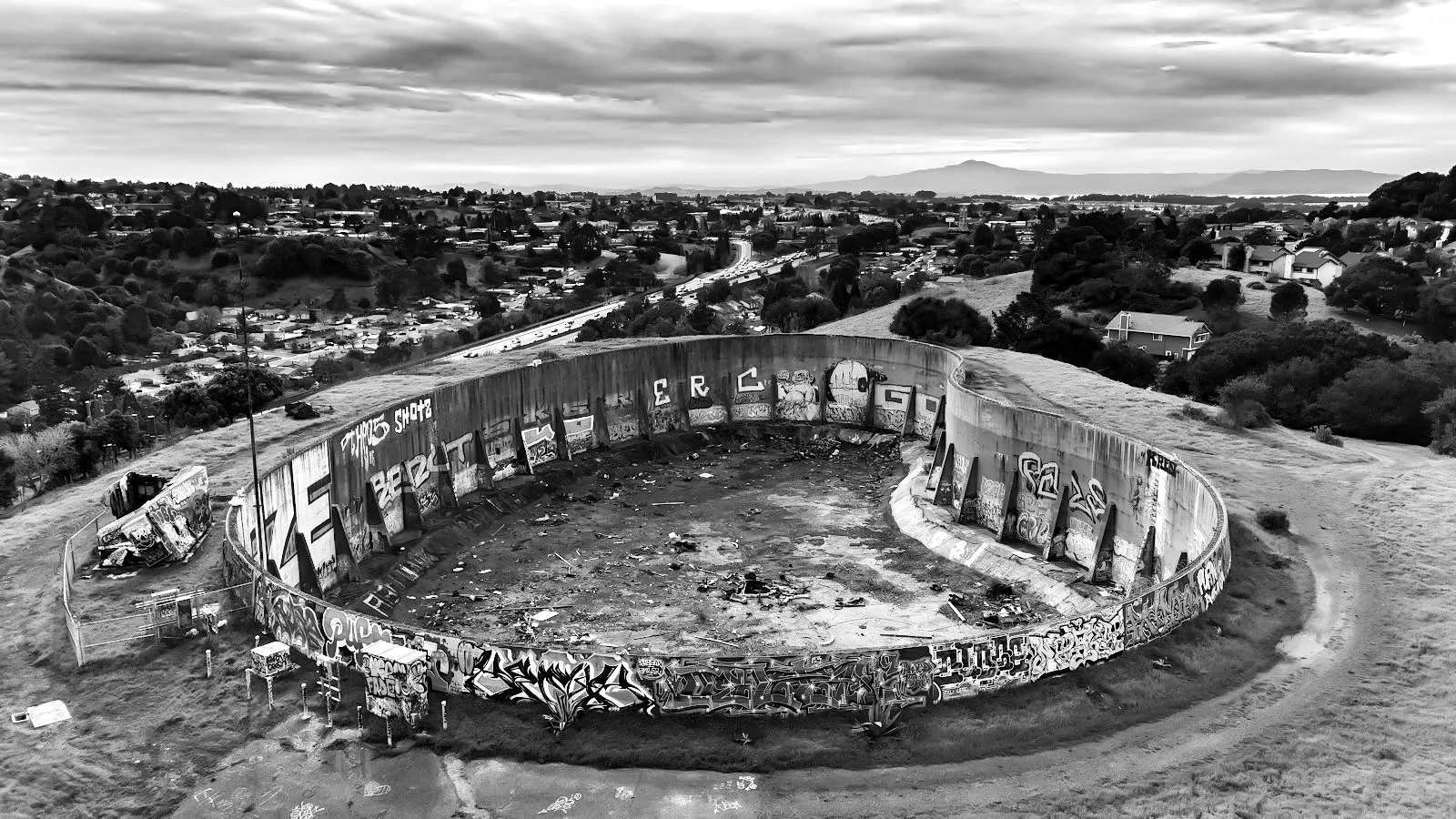 An empty, graffiti-covered concrete ruin on a hilltop overlooking a cityscape on an overcast day.