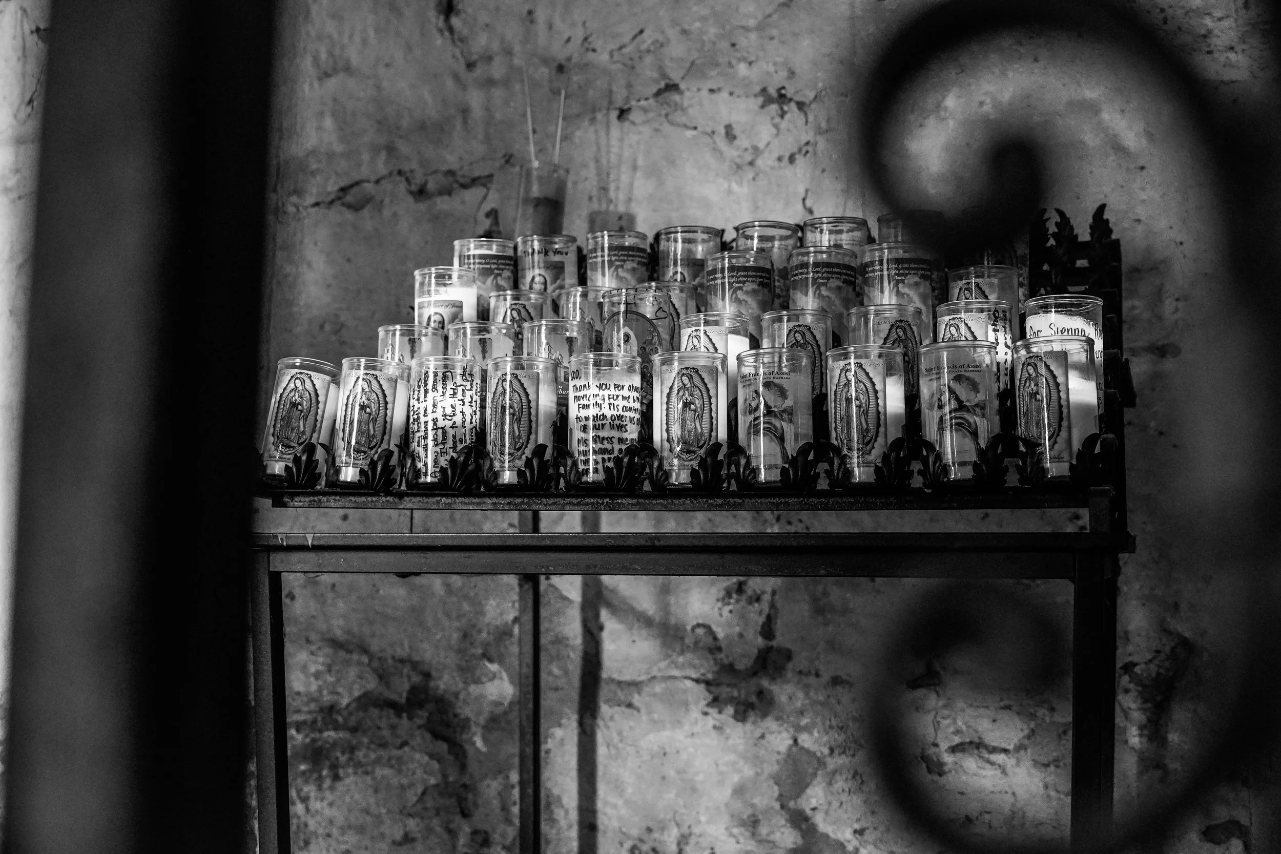 Black and white photo of a shelf filled with lit candles featuring religious images and notes, against a weathered stone wall, seen through a wrought iron gate.