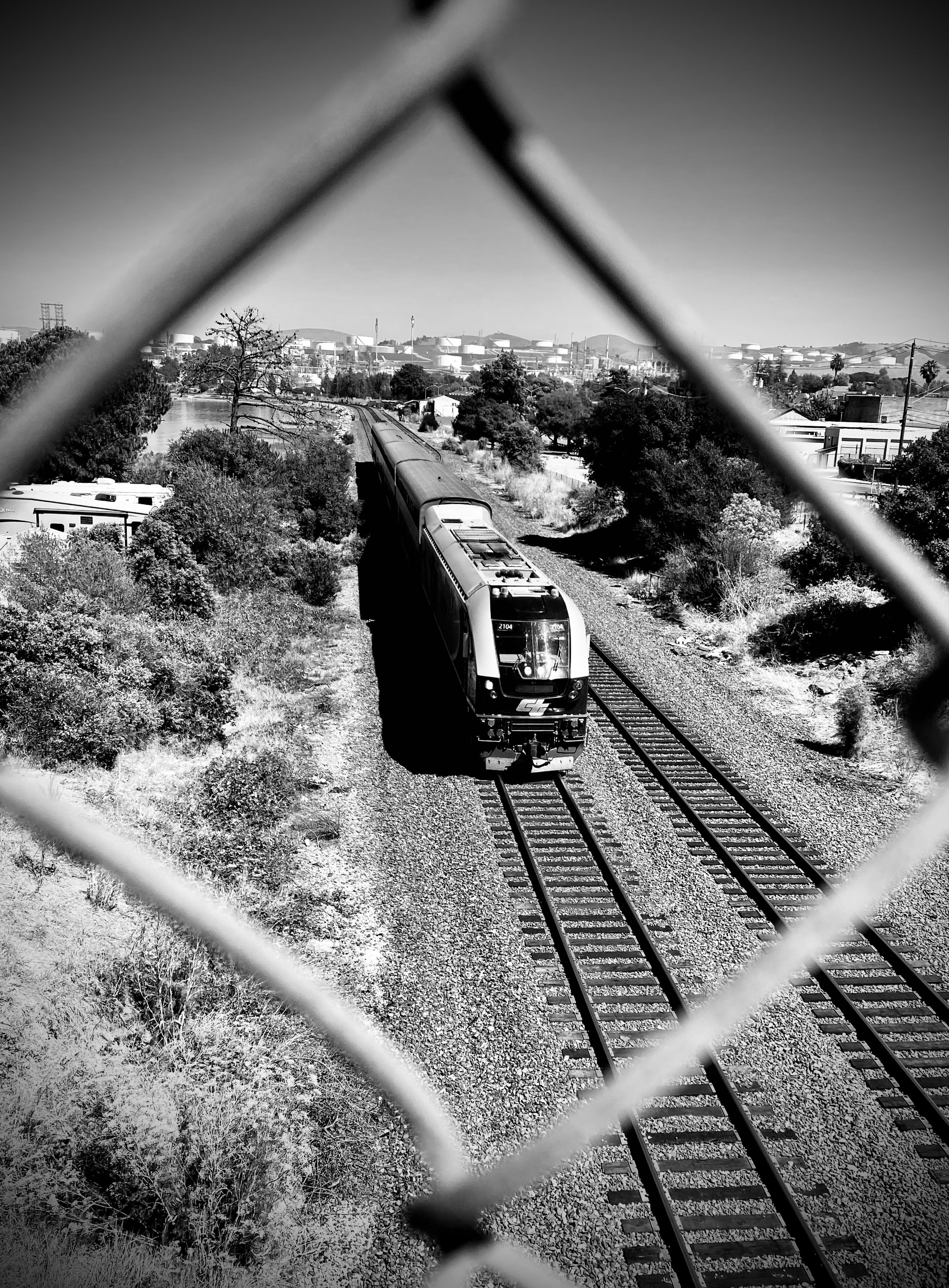 Black and white photo of a train moving along railway tracks, viewed through a chain-link fence.
