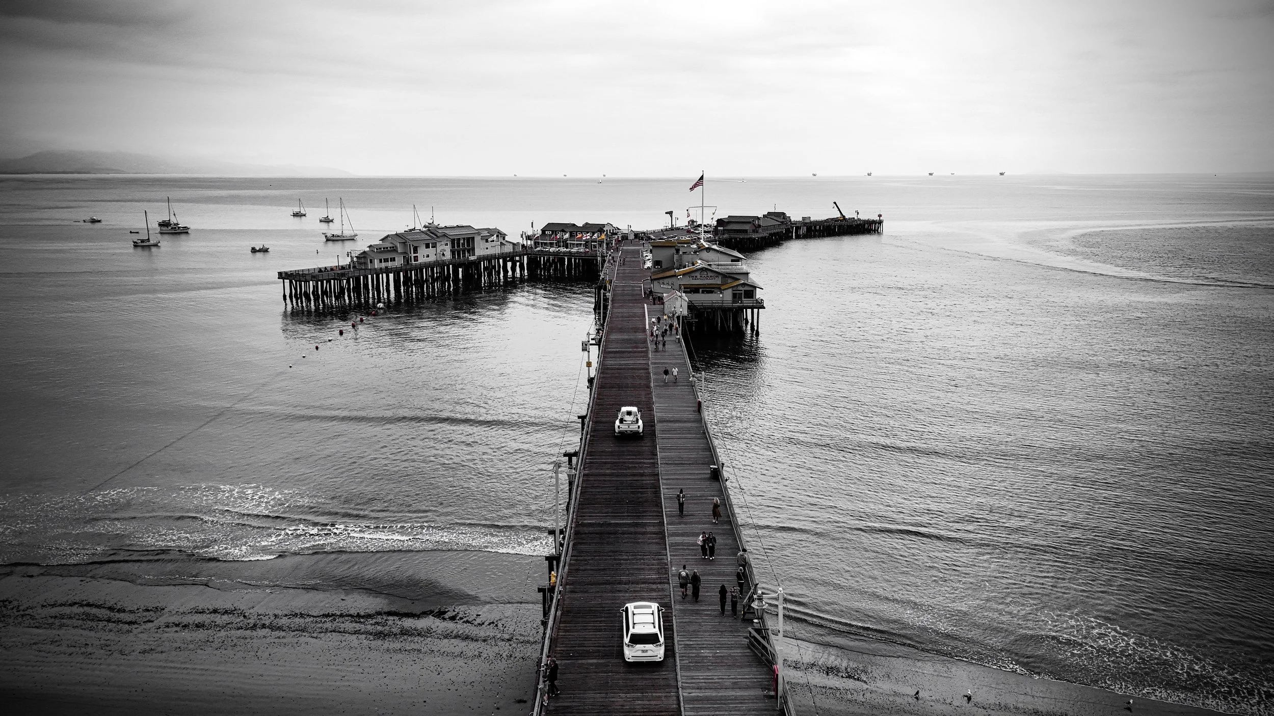 A black-and-white photo of a pier extending into the ocean with boats anchored nearby and people walking on the pier.