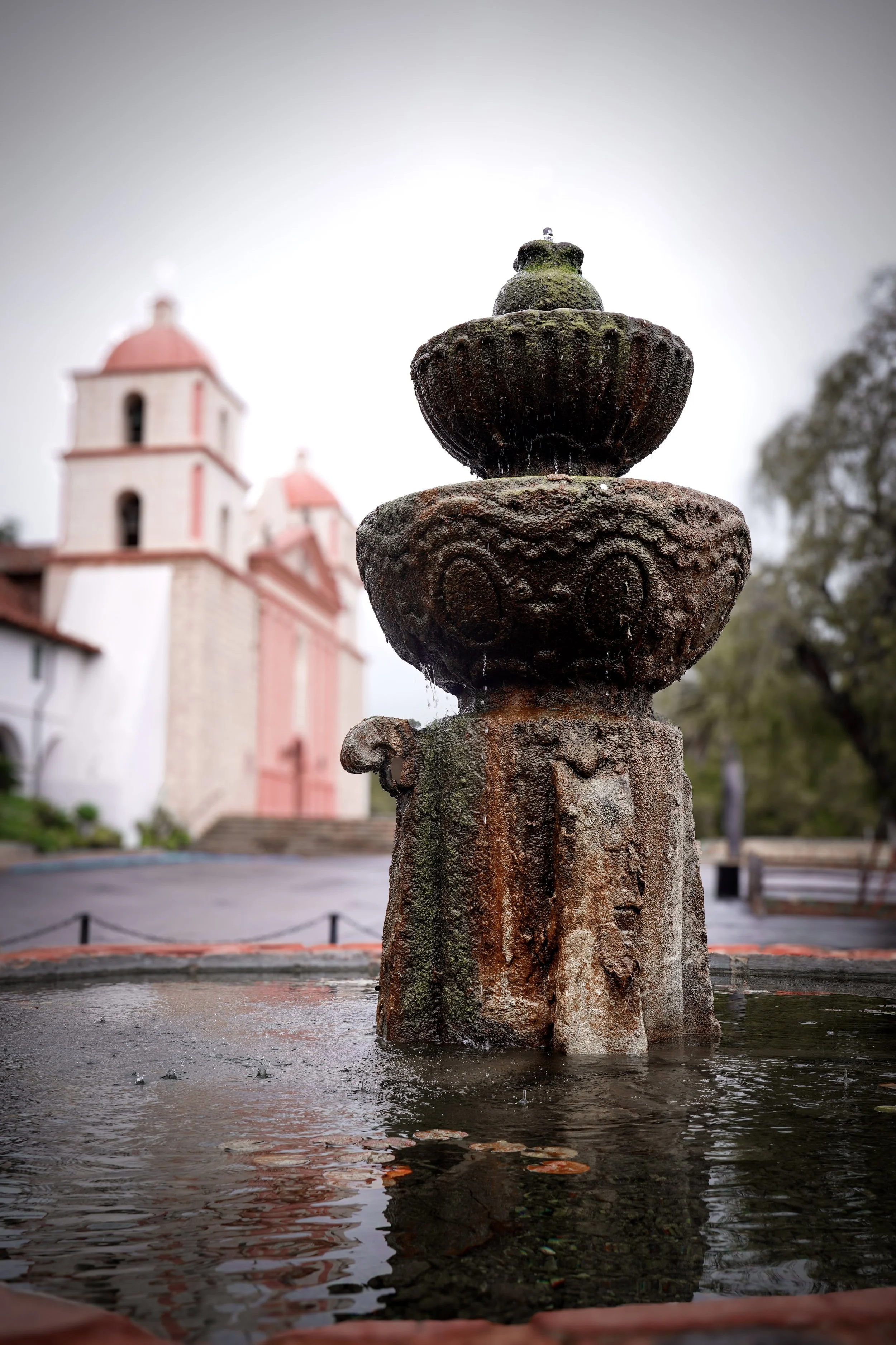 An old, moss-covered fountain with a decorative top in front of a church with pink and white walls and a red-tiled roof.