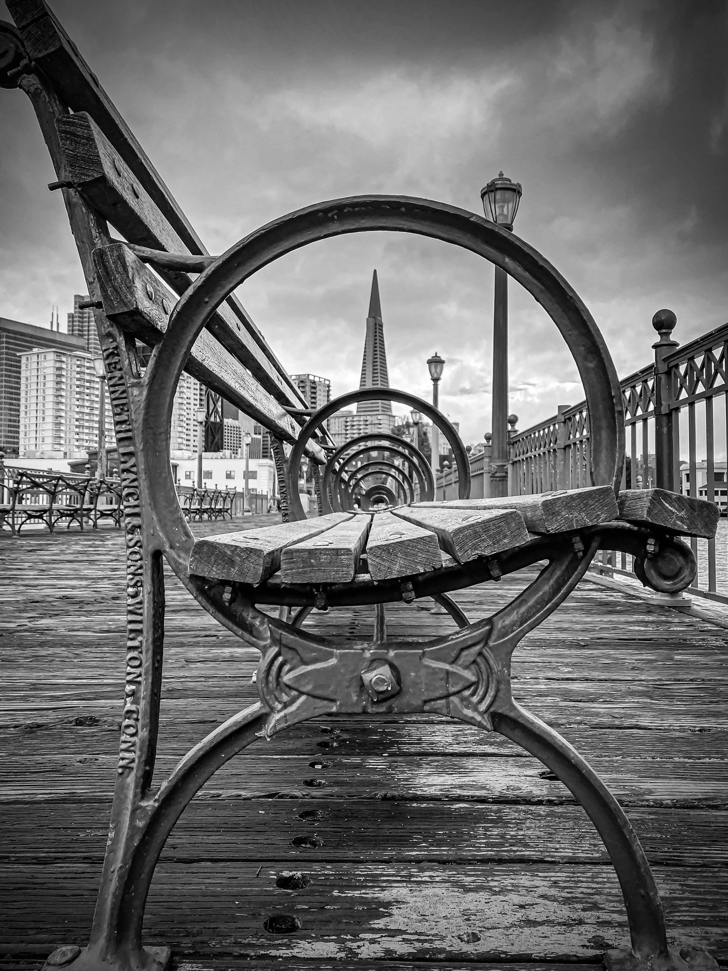 Black and white photo of a wooden park bench with decorative metal armrests and backrest, on a wooden boardwalk, with city skyscrapers and the Salesforce Tower in San Francisco in the background.