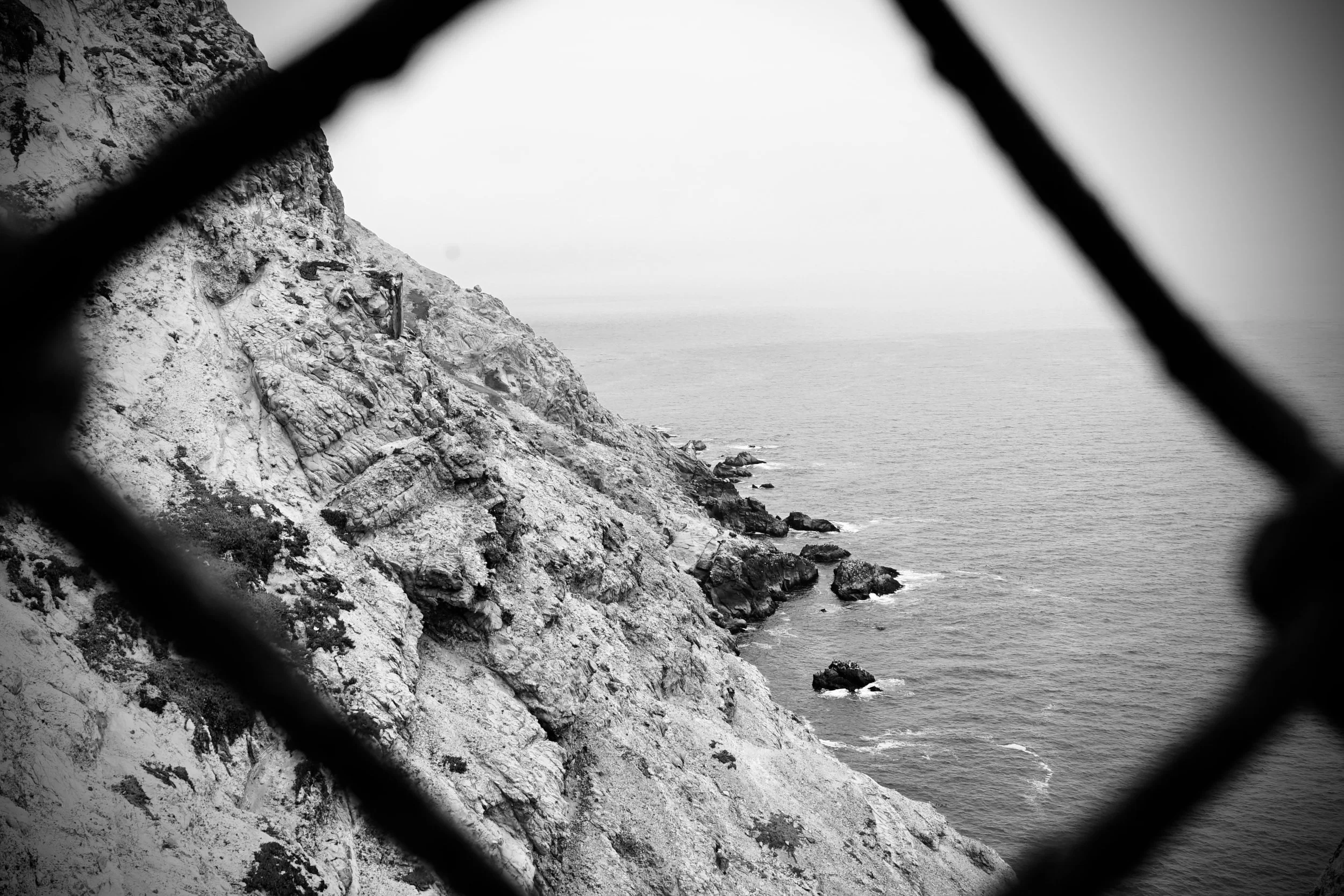 A black and white photo of a rocky coastal cliff seen through a chain-link fence.