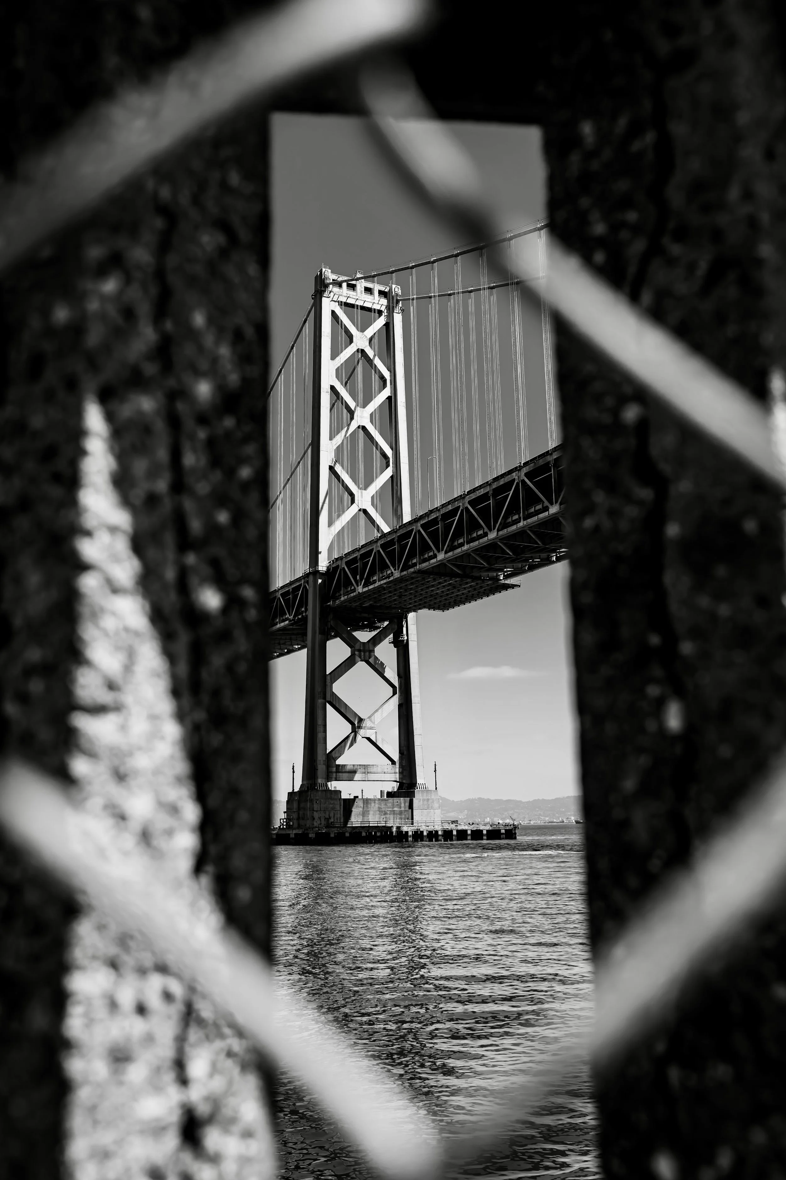 Black and white photo of a suspension bridge viewed through a small opening in a fence or grate.