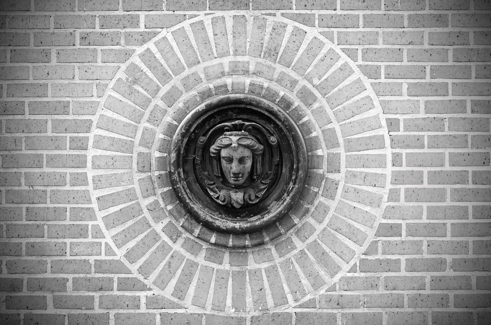 Close-up of a decorative wall medallion with a woman's face and hair detail, surrounded by brickwork arranged in a circular pattern.