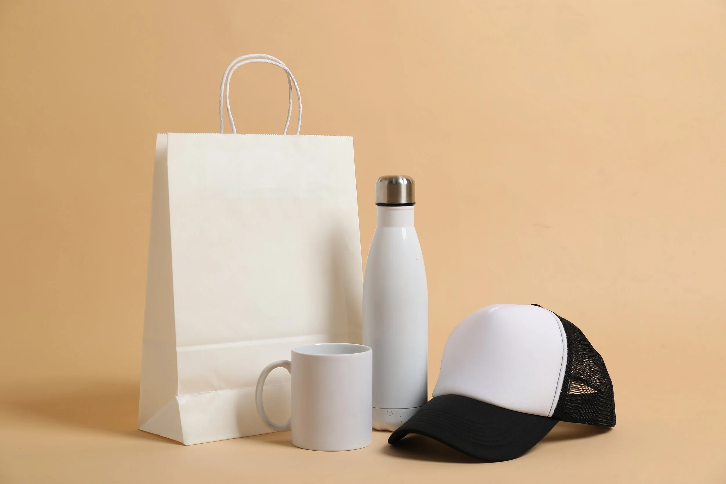 A collection of white and black objects on a beige background, including a paper bag, a mug, a water bottle, and a baseball cap.