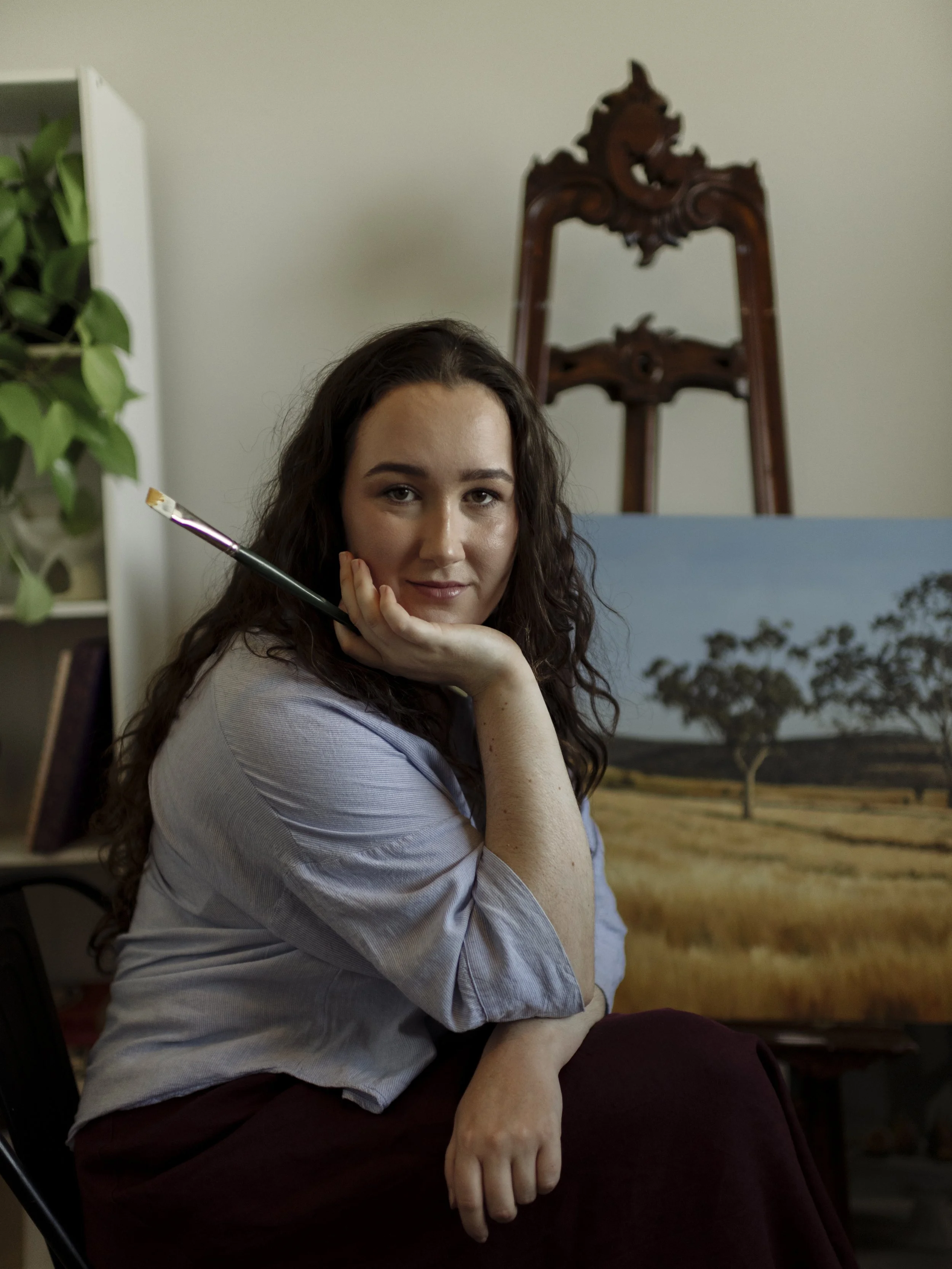 A portrait photo of artist Isabella Curtis in her home studio infront of a large wooden canvas and landscape painting