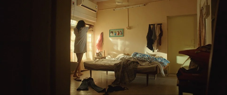 A woman stands on her toes, adjusting an air conditioning unit in a bedroom. The room has a bed with rumpled bedding, clothes hanging on the wall, and sunlight coming through the window.