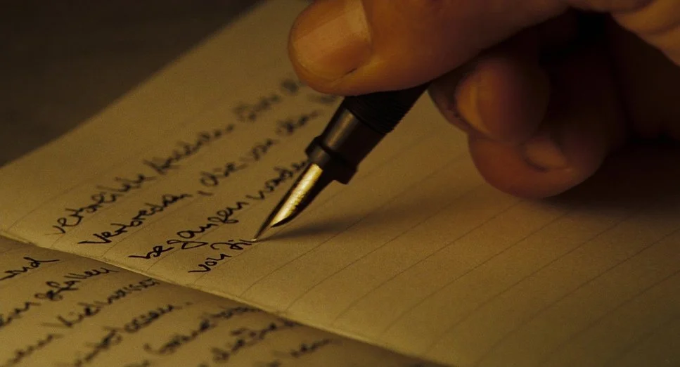 Close-up of a hand holding a black pen, writing on yellow lined paper filled with handwritten notes.