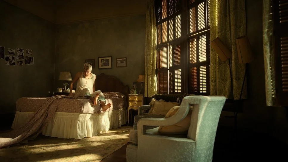 A woman sitting on a bed in a warmly lit bedroom, with sunlight streaming through the window blinds and yellow patterned curtains. The room has vintage furniture and framed photographs on the wall.