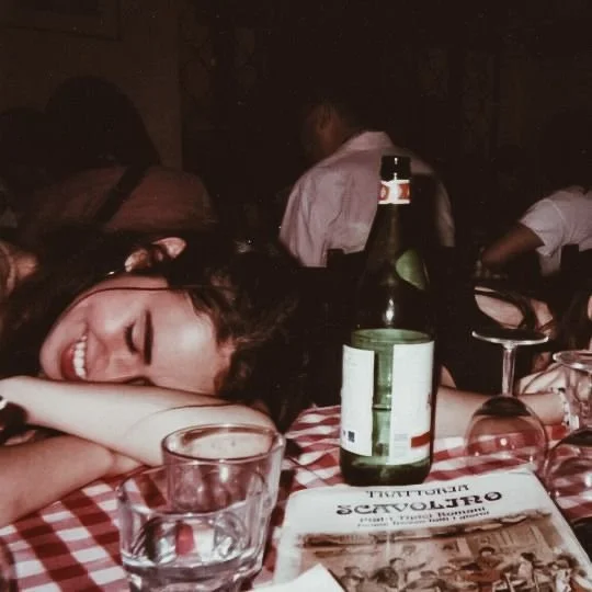 A woman smiling with eyes closed resting her head on her arms at a table with a checkered tablecloth, in a dimly lit restaurant or bar setting, with bottles, glasses, and a newspaper on the table.