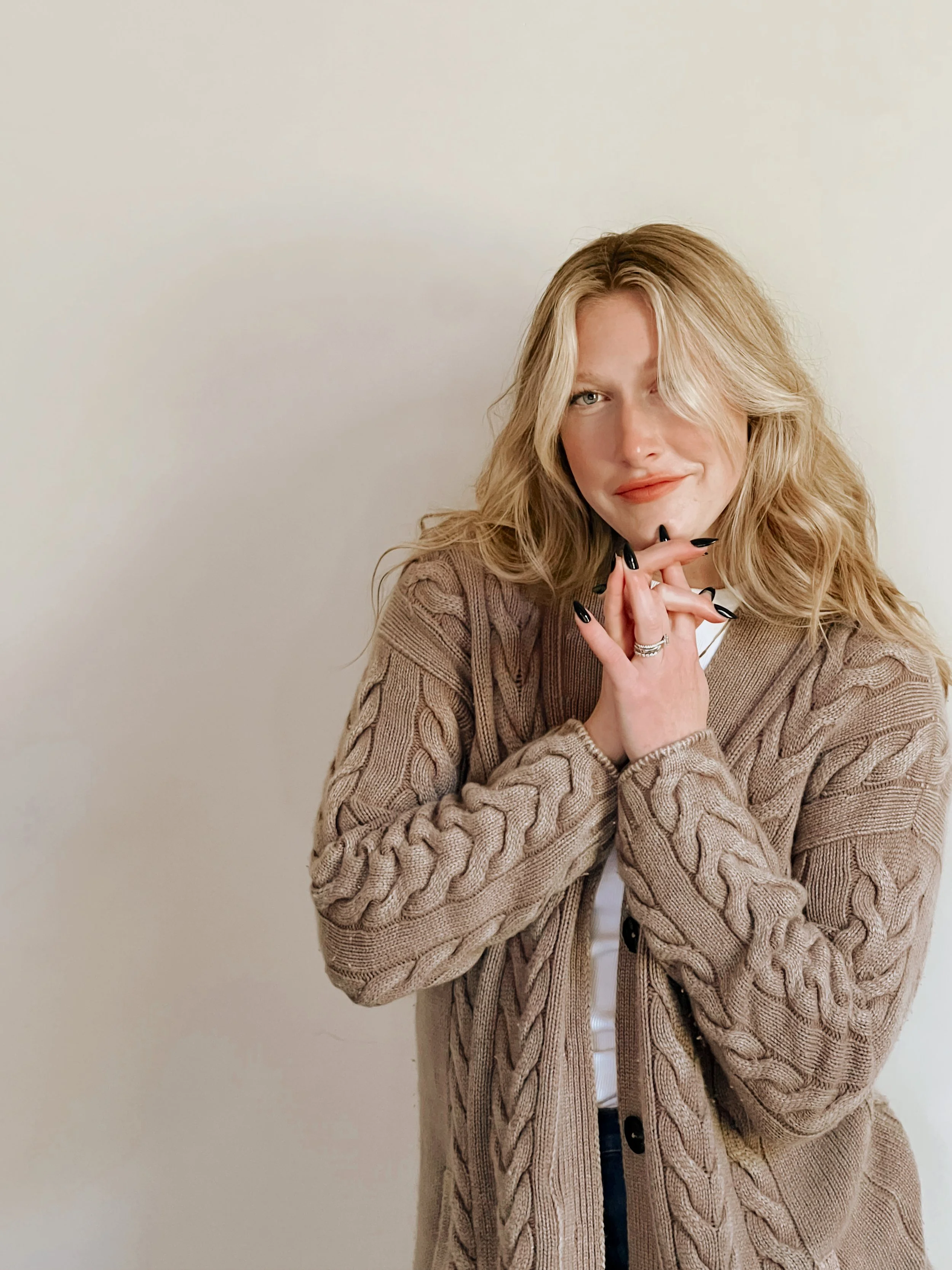 A woman with blonde wavy hair wearing a beige cable-knit cardigan and black nail polish, with her hands clasped near her chin, standing against a plain light-colored wall.
