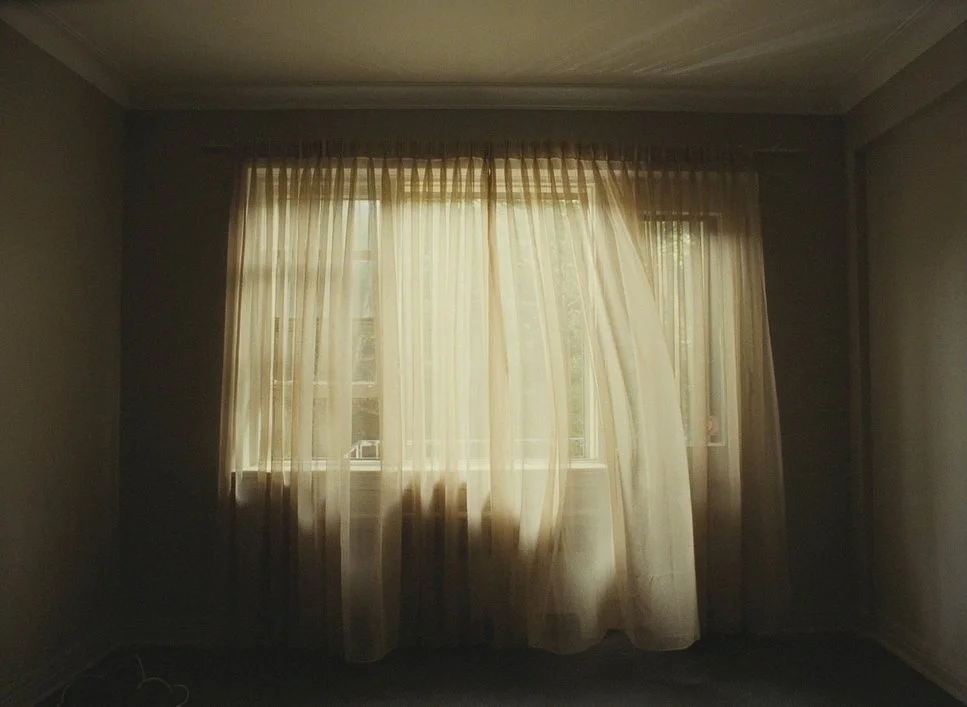 A window with sheer cream-colored curtains partly drawn, allowing sunlight to illuminate the room.