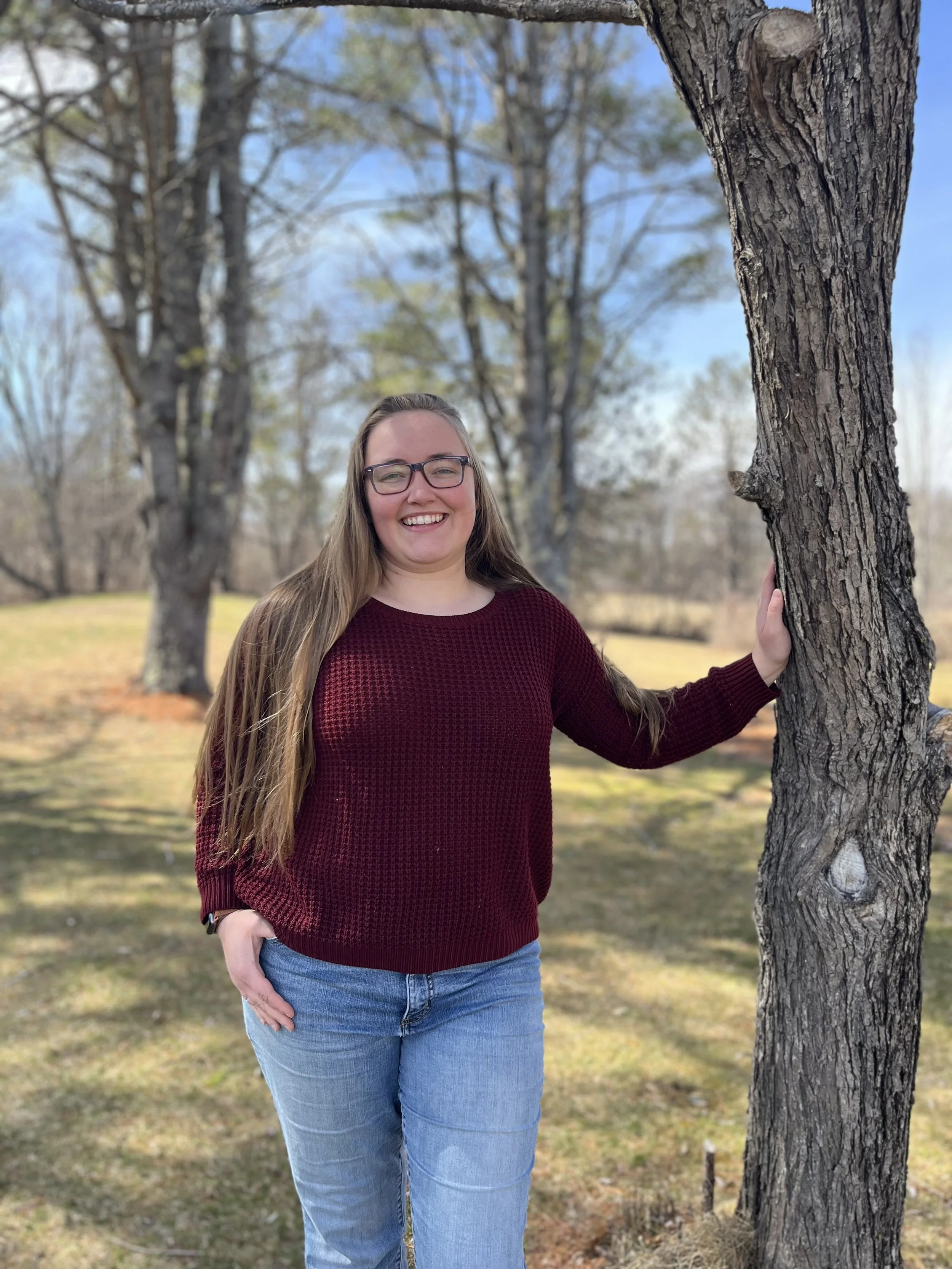 A woman with long hair, glasses, wearing a dark red sweater and blue jeans, standing outdoors near a tree with a grassy background and blue sky.