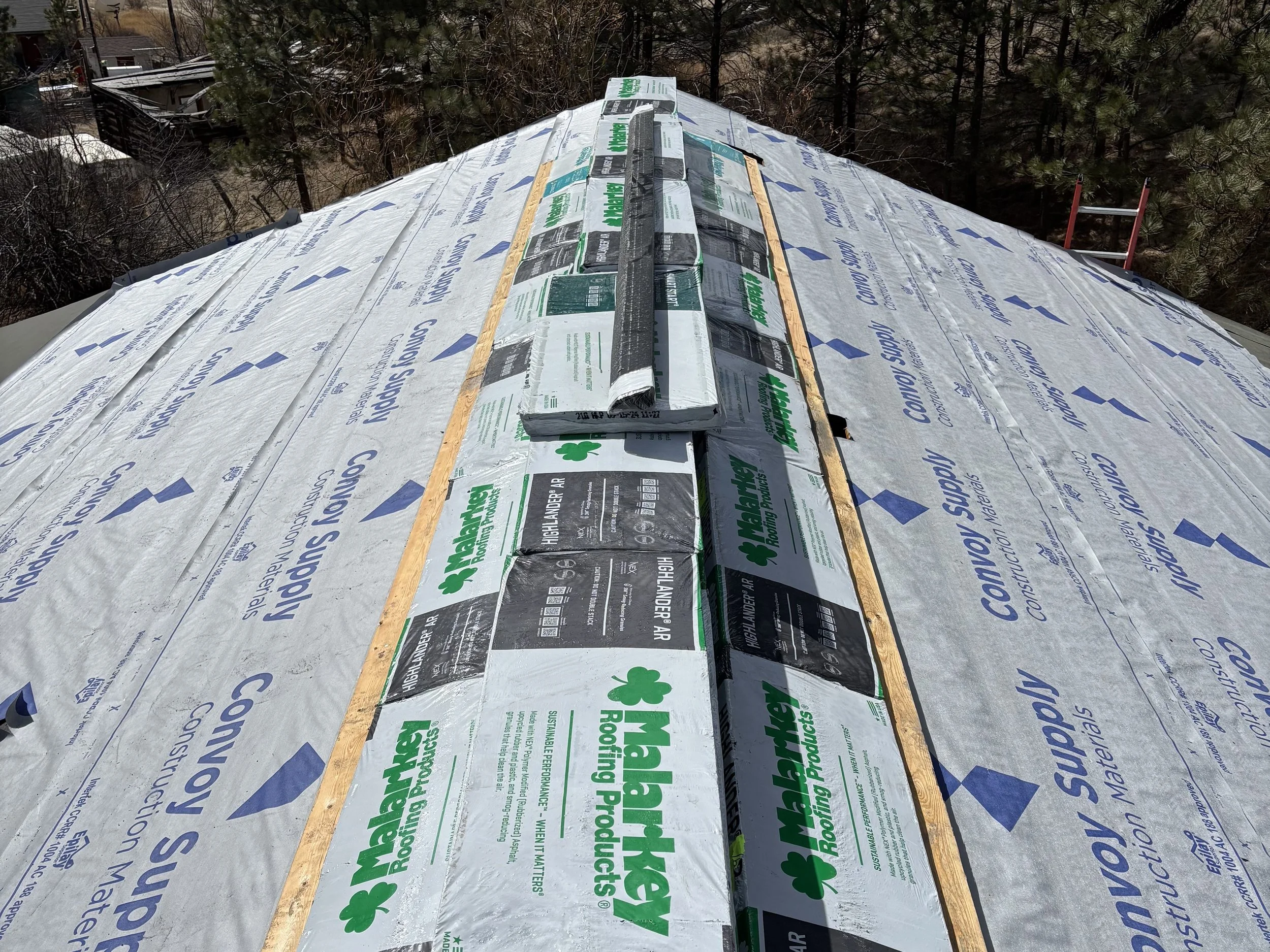 Roof under construction with roofing underlayment and shingles stacked in the center, surrounded by trees.