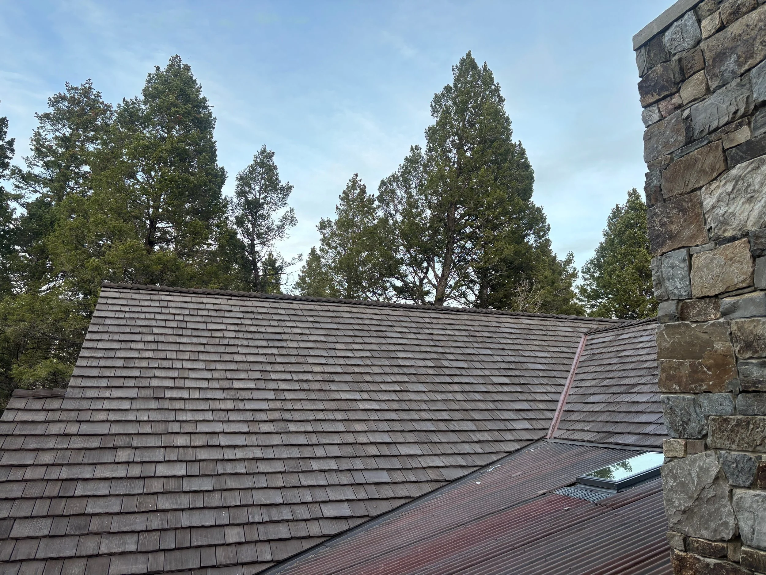 View of a house's roof with brown shingles, a chimney on the right side made of stone, and a small skylight. Tall trees and a cloudy sky in the background.