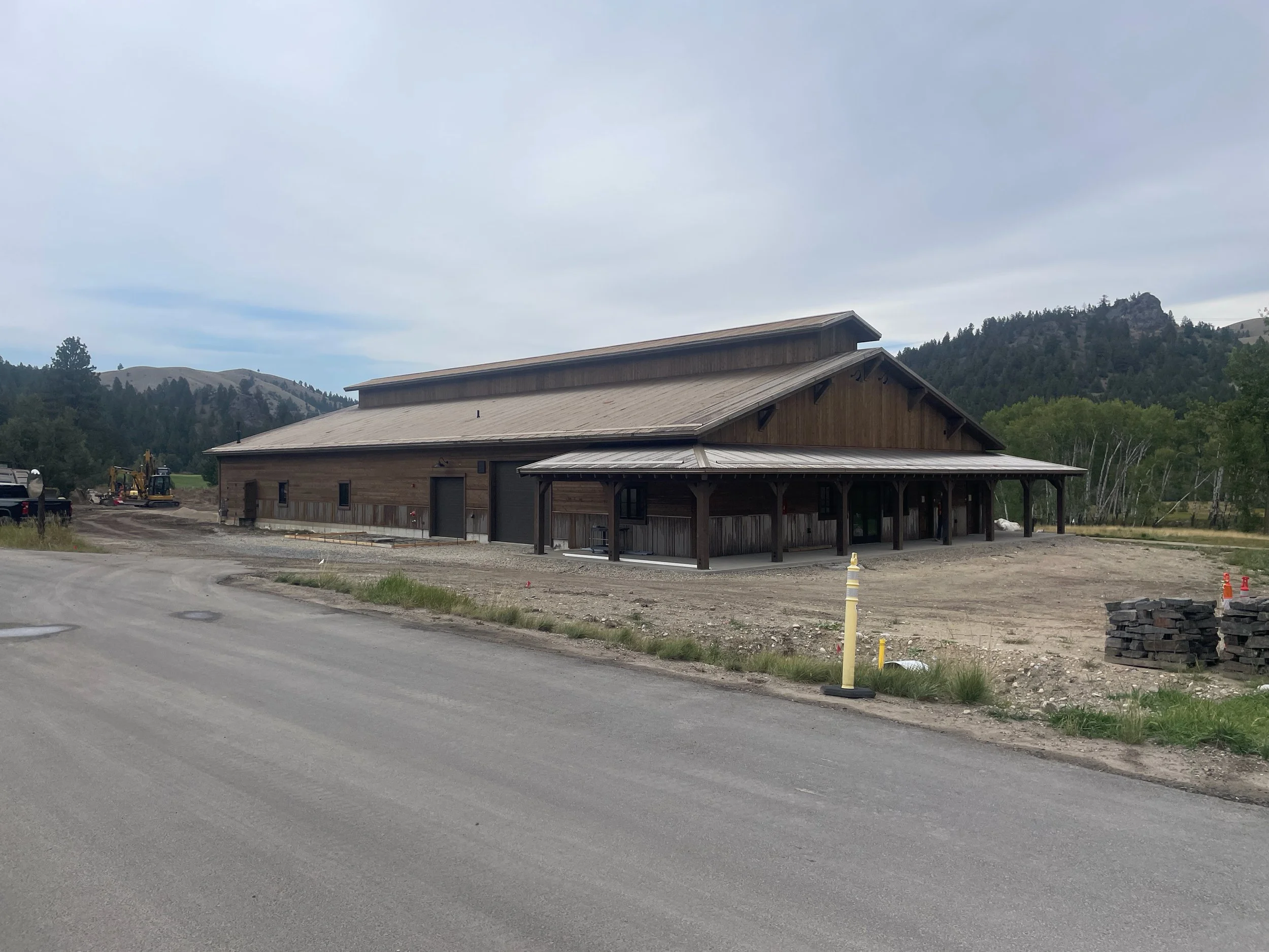 A large wooden barn under construction in a rural area surrounded by mountains and trees.