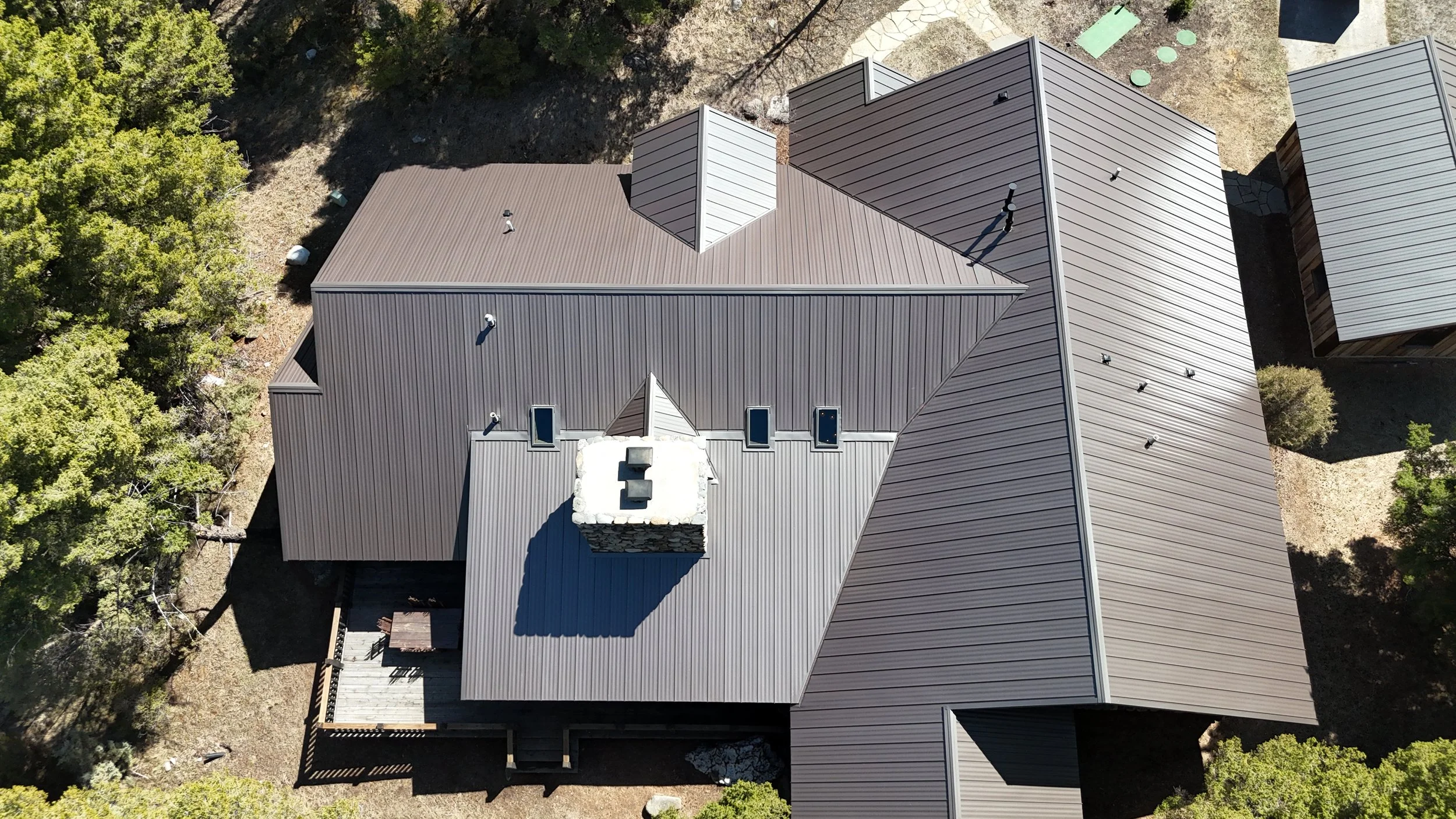 An aerial view of a house with a complex metal roof, surrounded by trees and a small deck with outdoor furniture.