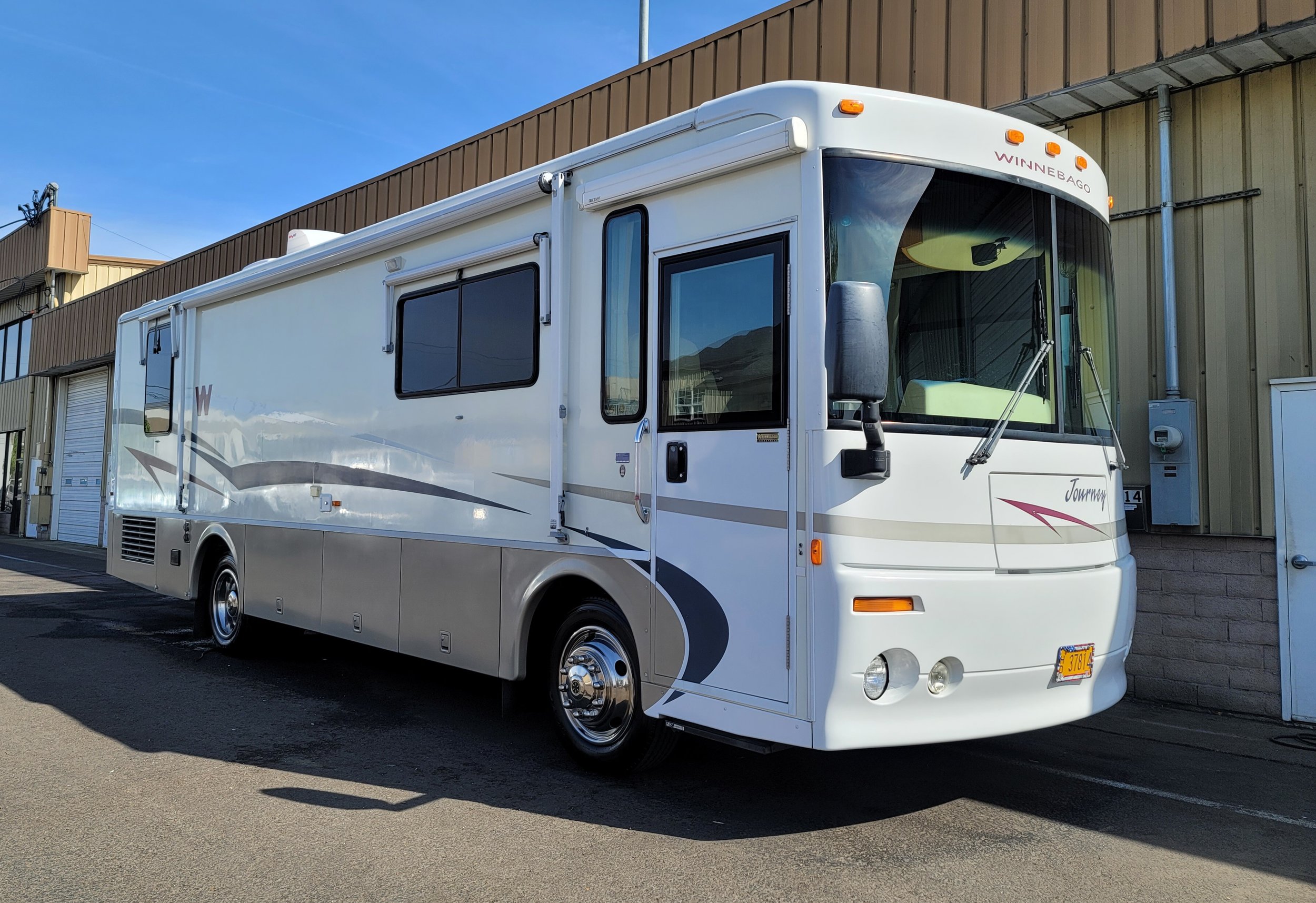 White Winnebago Journey motorhome parked outdoors next to a building with a blue sky overhead.