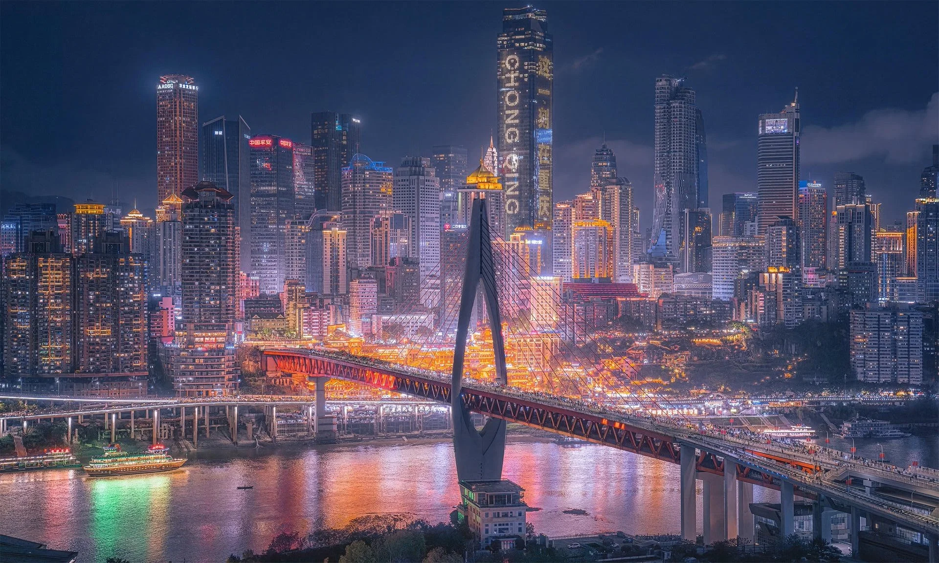 Night view of a city skyline with skyscrapers illuminated, a bridge over a river, and boats on the water, showcasing vibrant city lights.