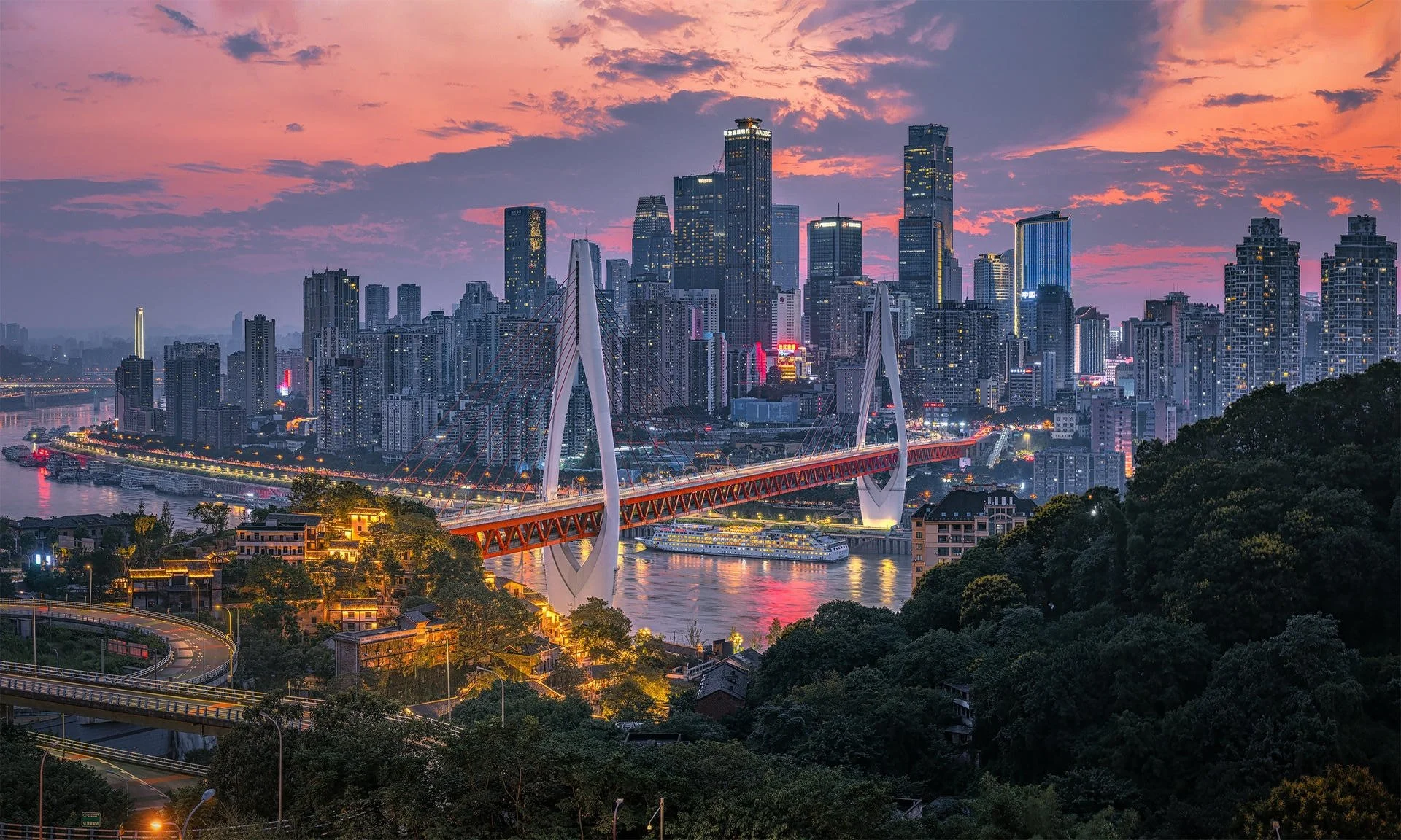 Skyline of a city at dusk with tall skyscrapers, a bridge spanning a river, and boats docked along the water, with pink and purple clouds in the sky.