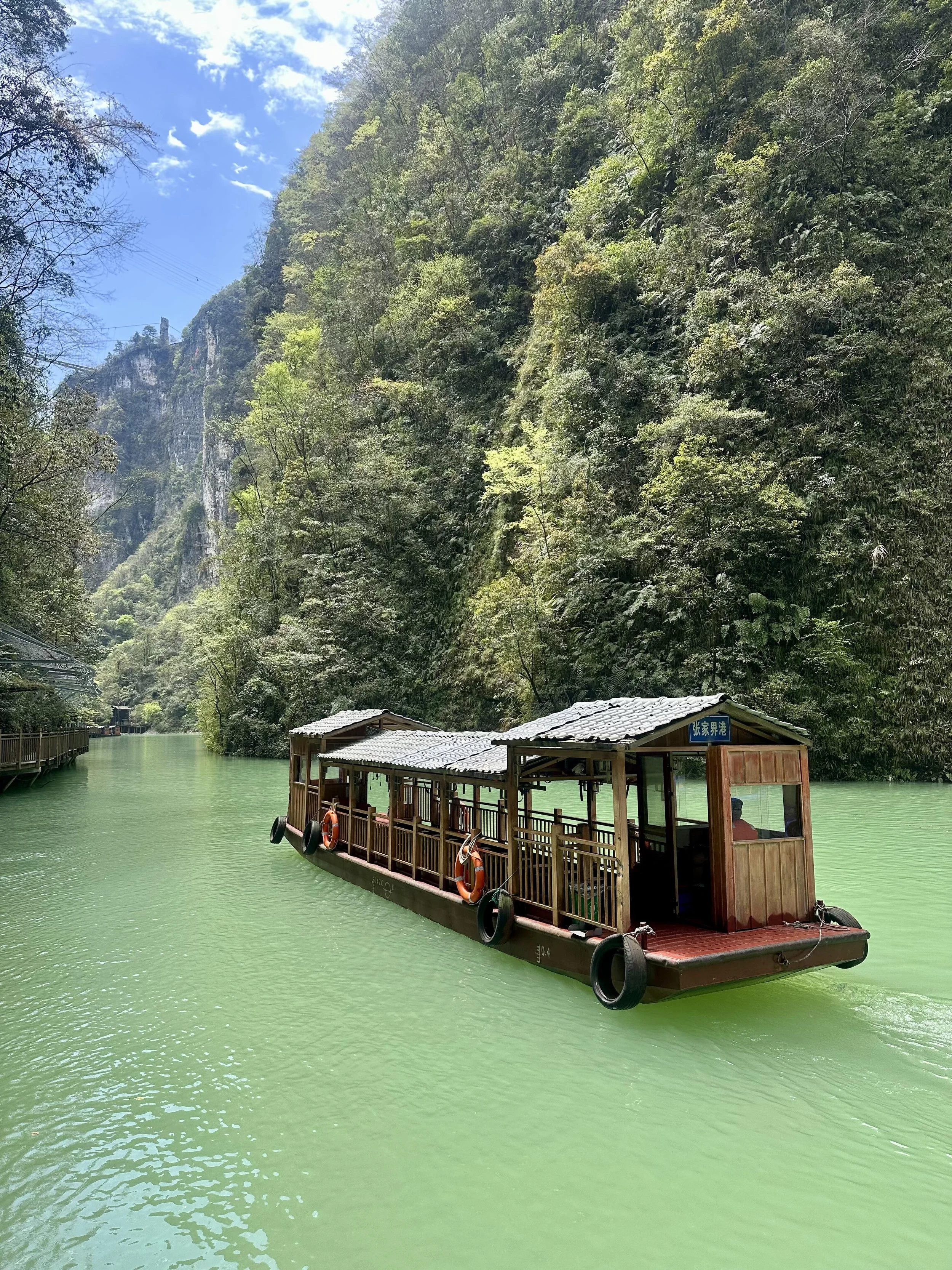 A wooden boat with a roof and orange life rings, floating on a green river surrounded by lush green forested mountains under a partly cloudy sky.