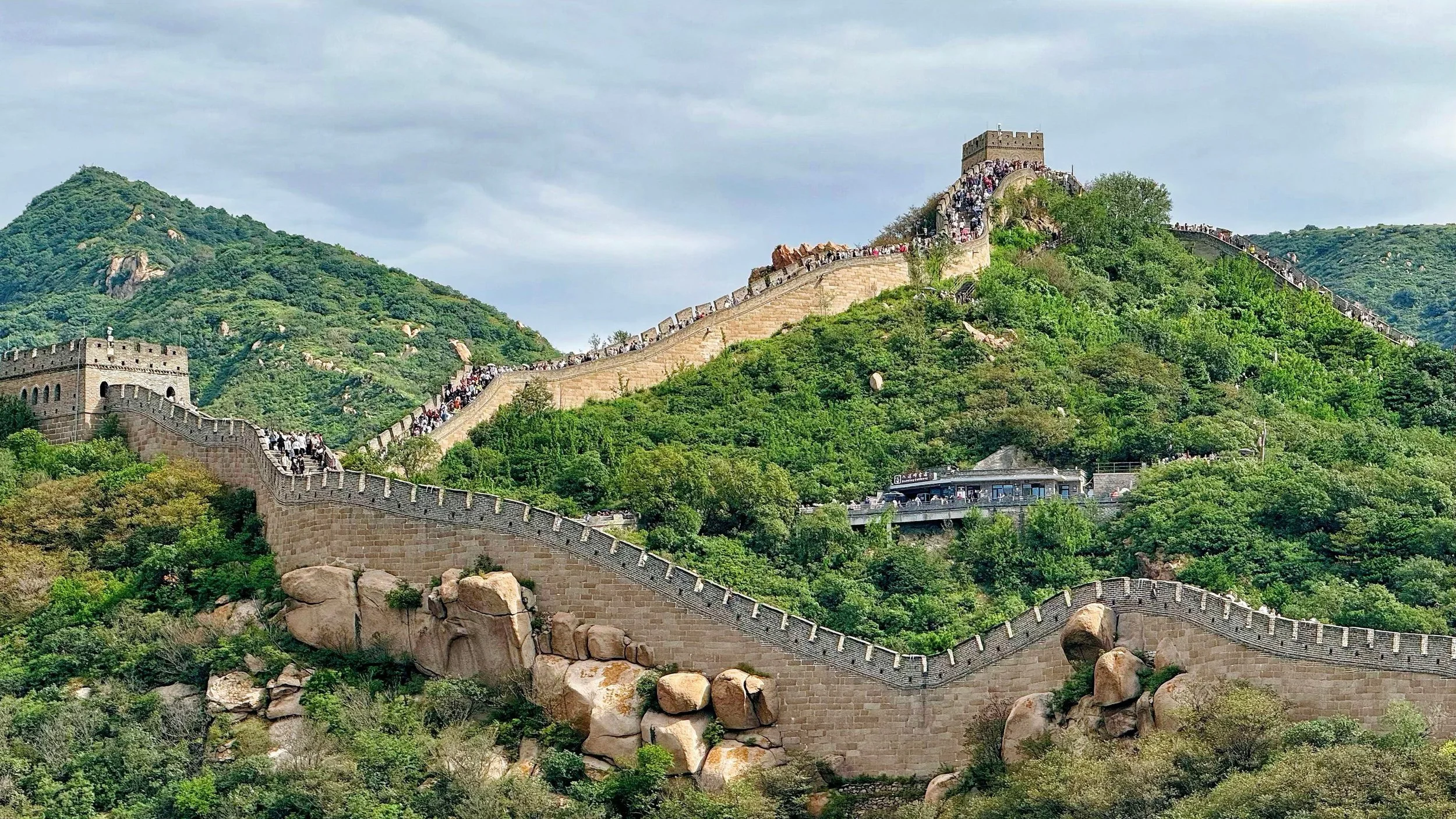 The Great Wall of China winding through green, hilly terrain with a building at the top and crowds of visitors walking along the wall.