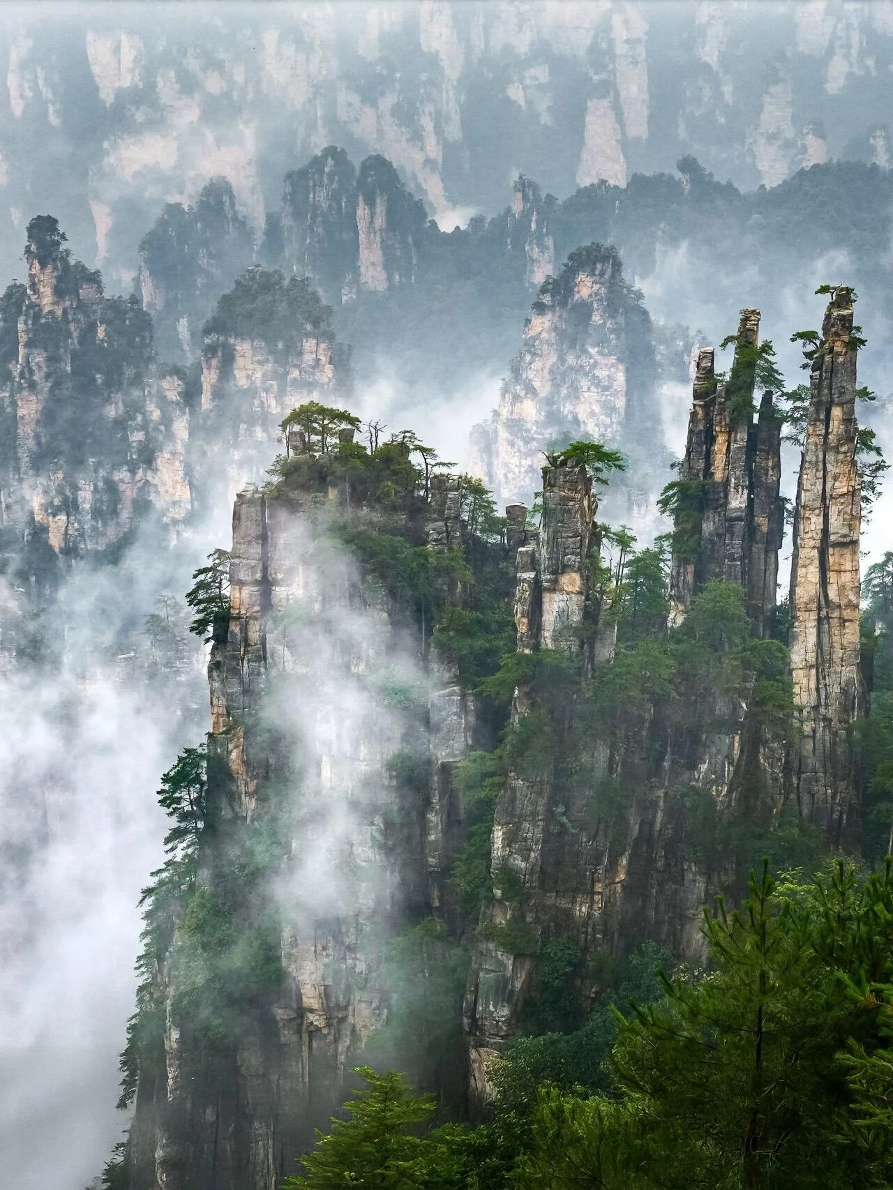 Tall, narrow rock formations covered with green trees and fog in a mountain landscape.
