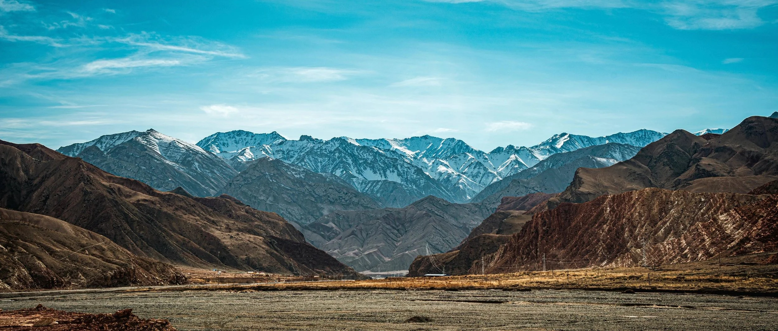 Mountain range with snow-capped peaks under a blue sky, rugged hills in the foreground.