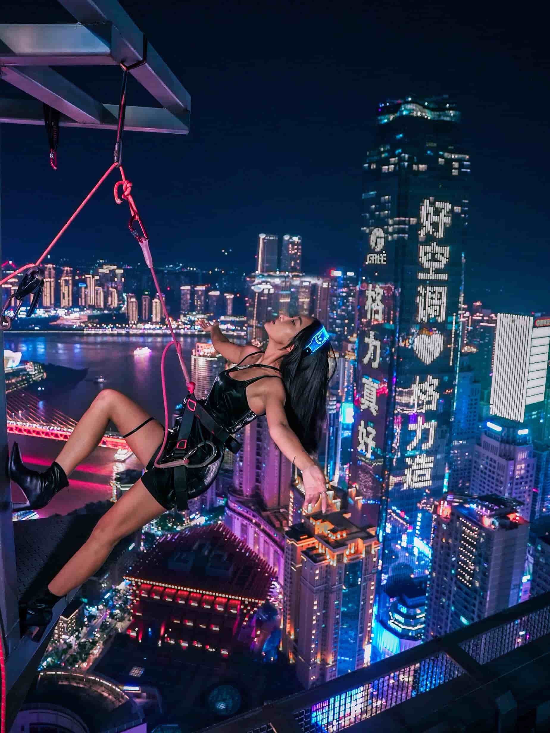 A woman sitting on a ledge overlooking a city skyline at night, with neon lights and tall buildings, some with illuminated signs, in the background. She is wearing a black outfit and headphones, with her head tilted back and arms outstretched.