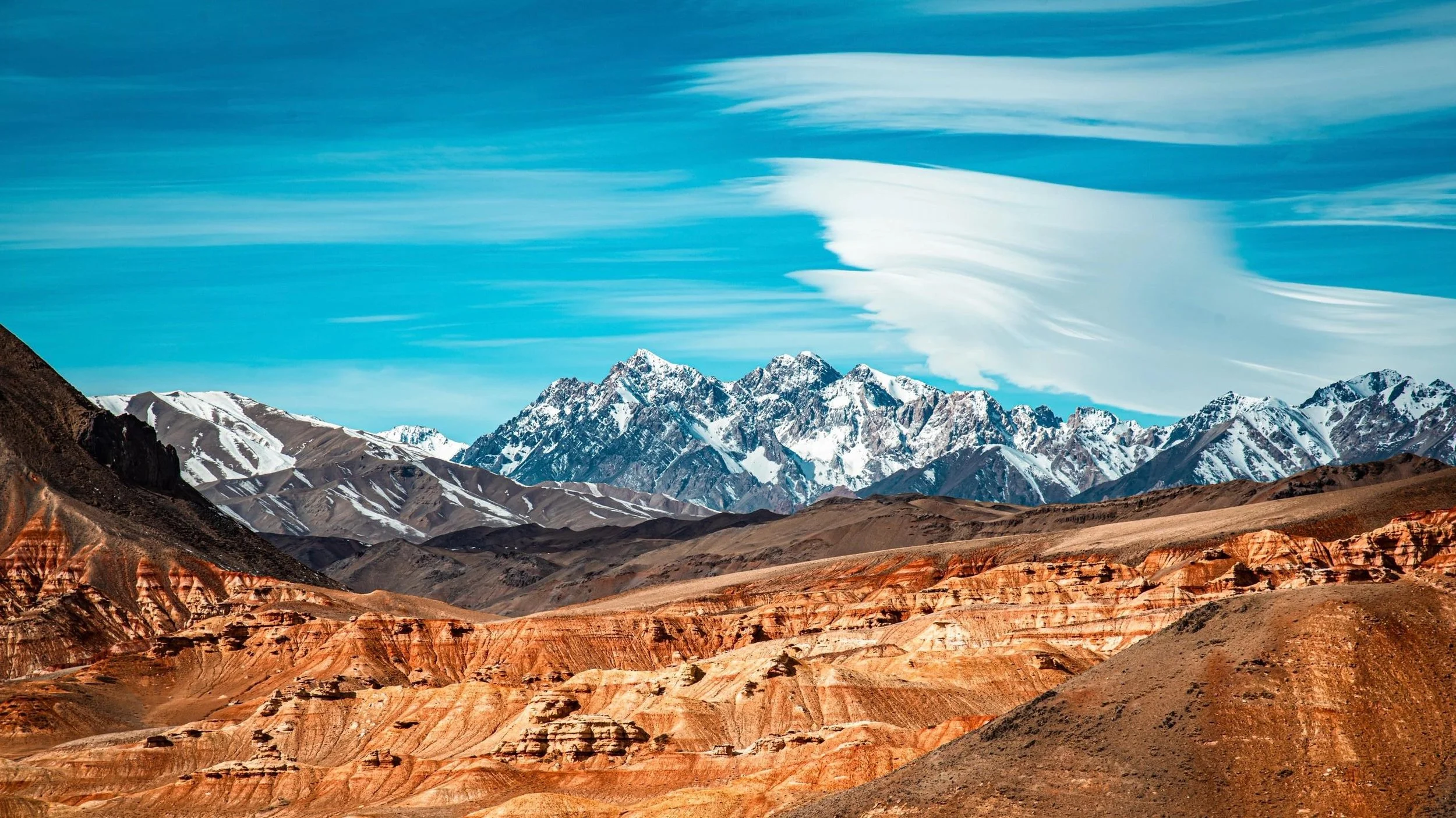 Snow-capped mountains under a blue sky with wispy clouds, arid desert terrain in the foreground.