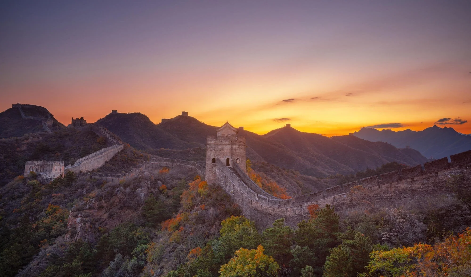 The Great Wall of China winding over hills at sunset with colorful trees in the foreground.