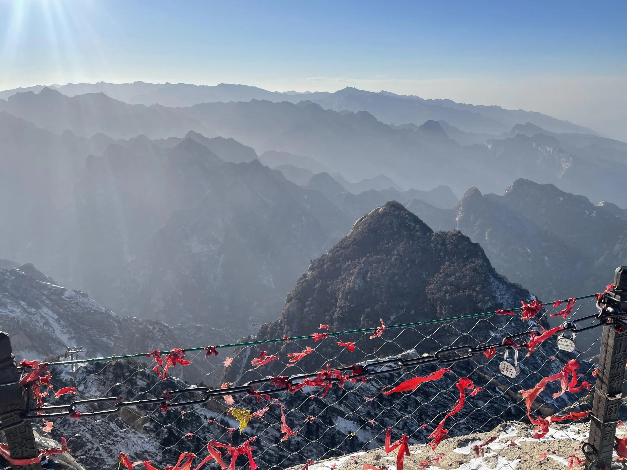Mountain landscape with layers of mountains extending into the distance, under a clear blue sky with sunlight shining from the top left corner. In the foreground, there is a safety net with red ribbons attached, secured to a viewing platform with a mechanical device on the right side.