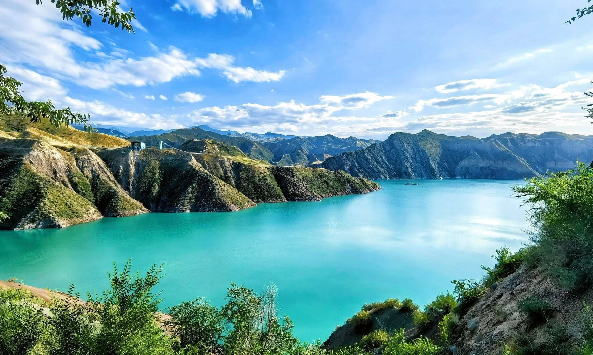 Scenic view of a turquoise lake surrounded by green hills and mountains under a partly cloudy blue sky, with foliage in the foreground.