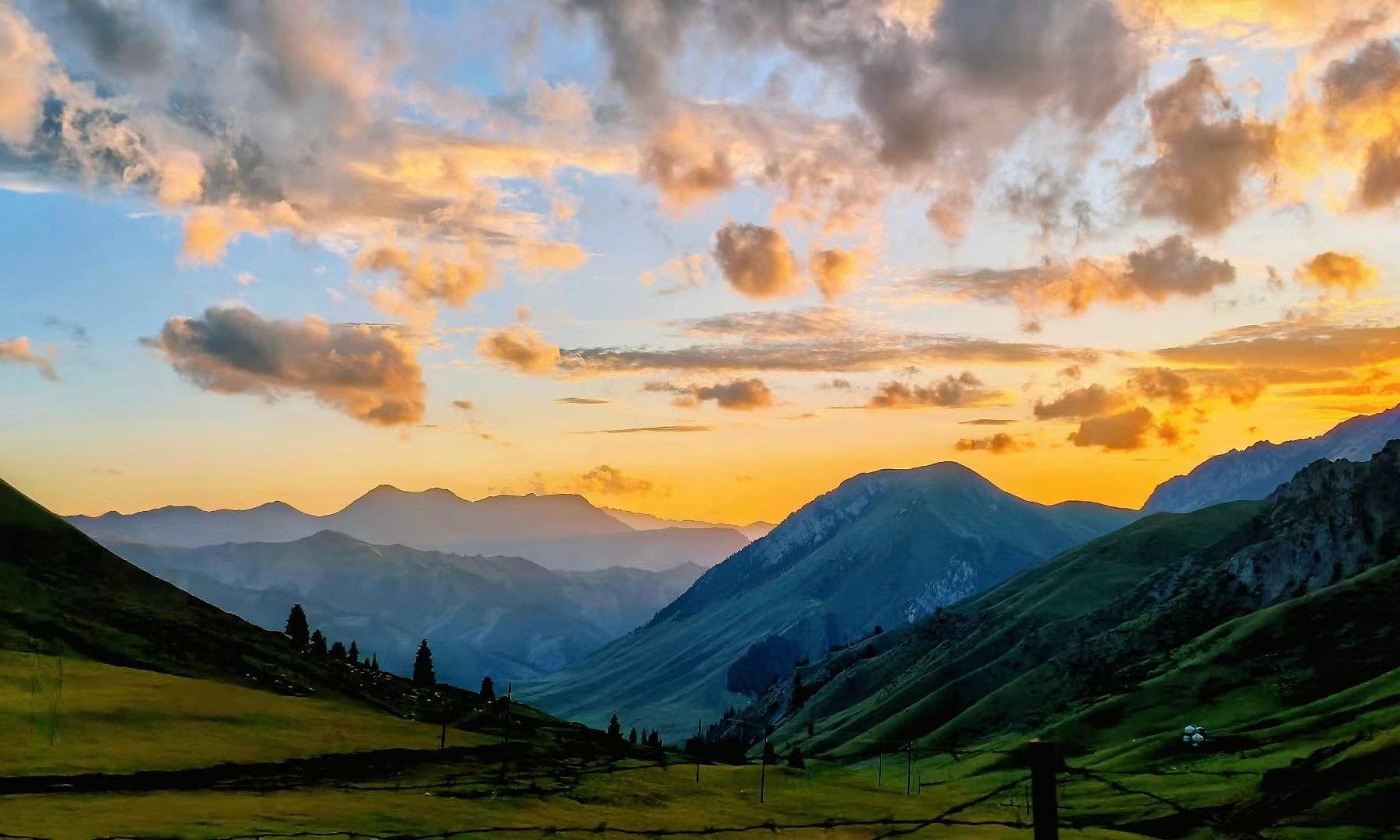 A panoramic view of mountains during sunset with a partly cloudy sky and green hills in the foreground.