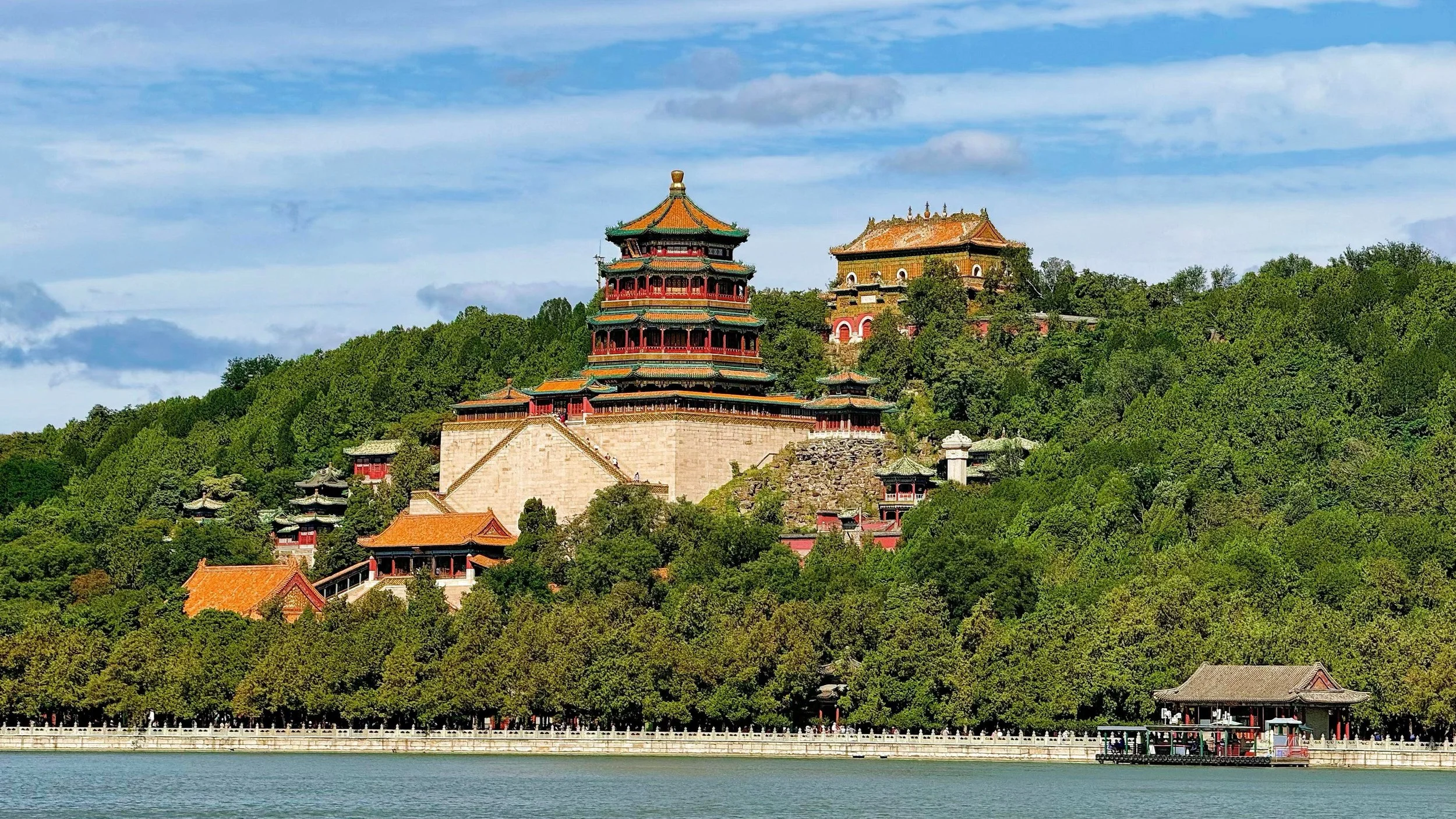 Traditional Chinese pagoda on a hill surrounded by trees, with a body of water in the foreground and a partly cloudy sky in the background.