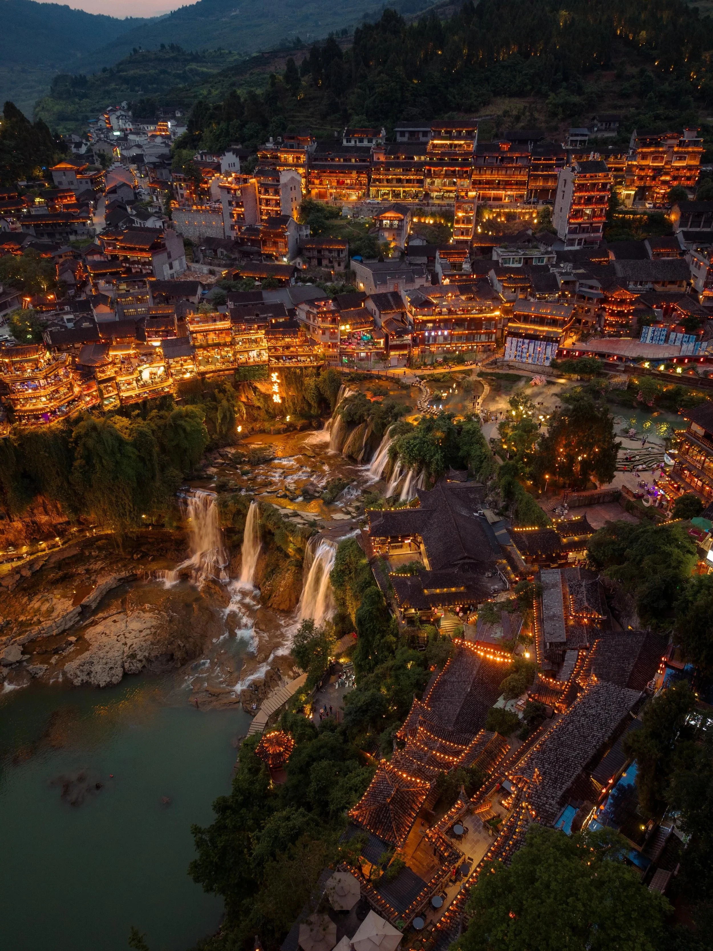 Aerial view of Furong Town ancient village at dusk — clusters of Tujia stilted houses on the cliff above the waterfall, Yongshun County, Hunan