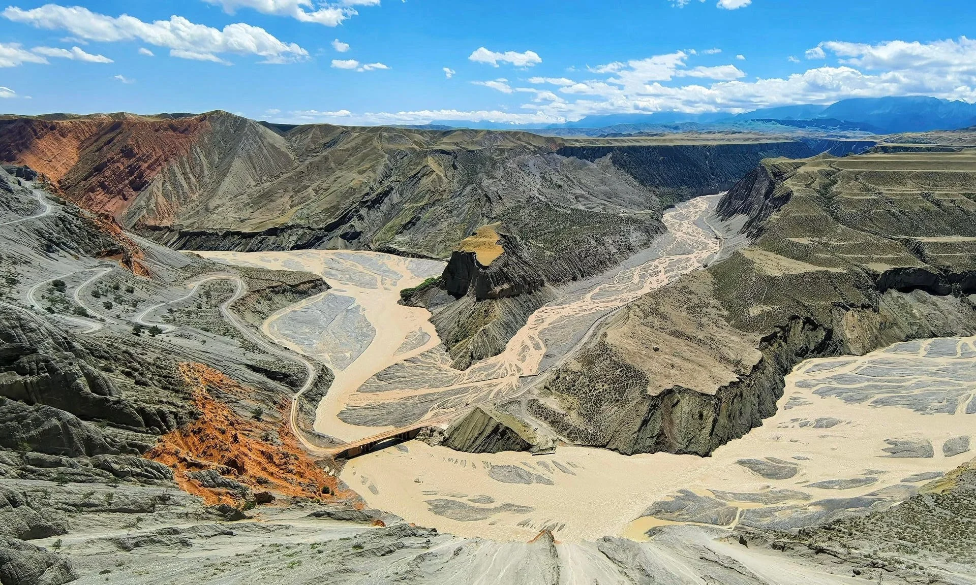 Aerial view of large canyon with winding dirt roads and braided rivers, mountains in the background under a partly cloudy sky.