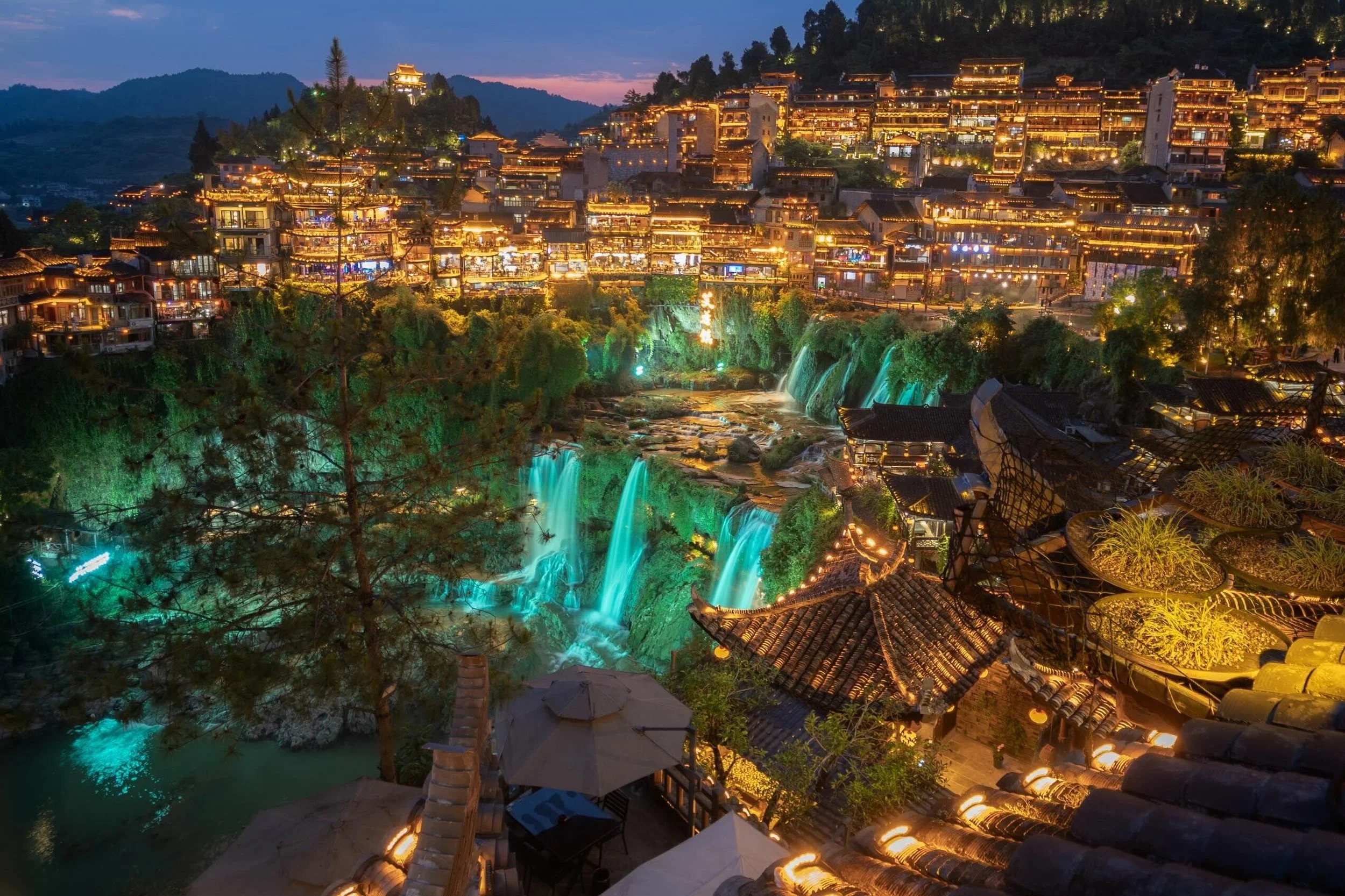 Turquoise waterfall flowing beneath Furong Town at blue hour — traditional temple roofs and wooden walkways along the falls, Hunan, China