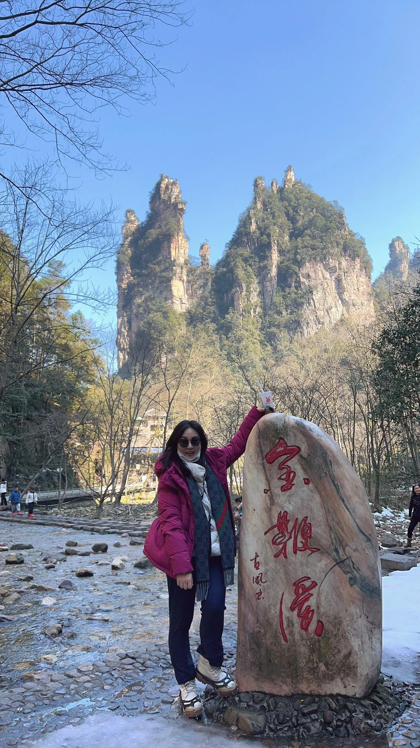 A woman wearing a pink jacket and sunglasses standing next to a large rock with red Chinese characters engraved on it. The landscape features tall mountains, trees, and a clear blue sky.