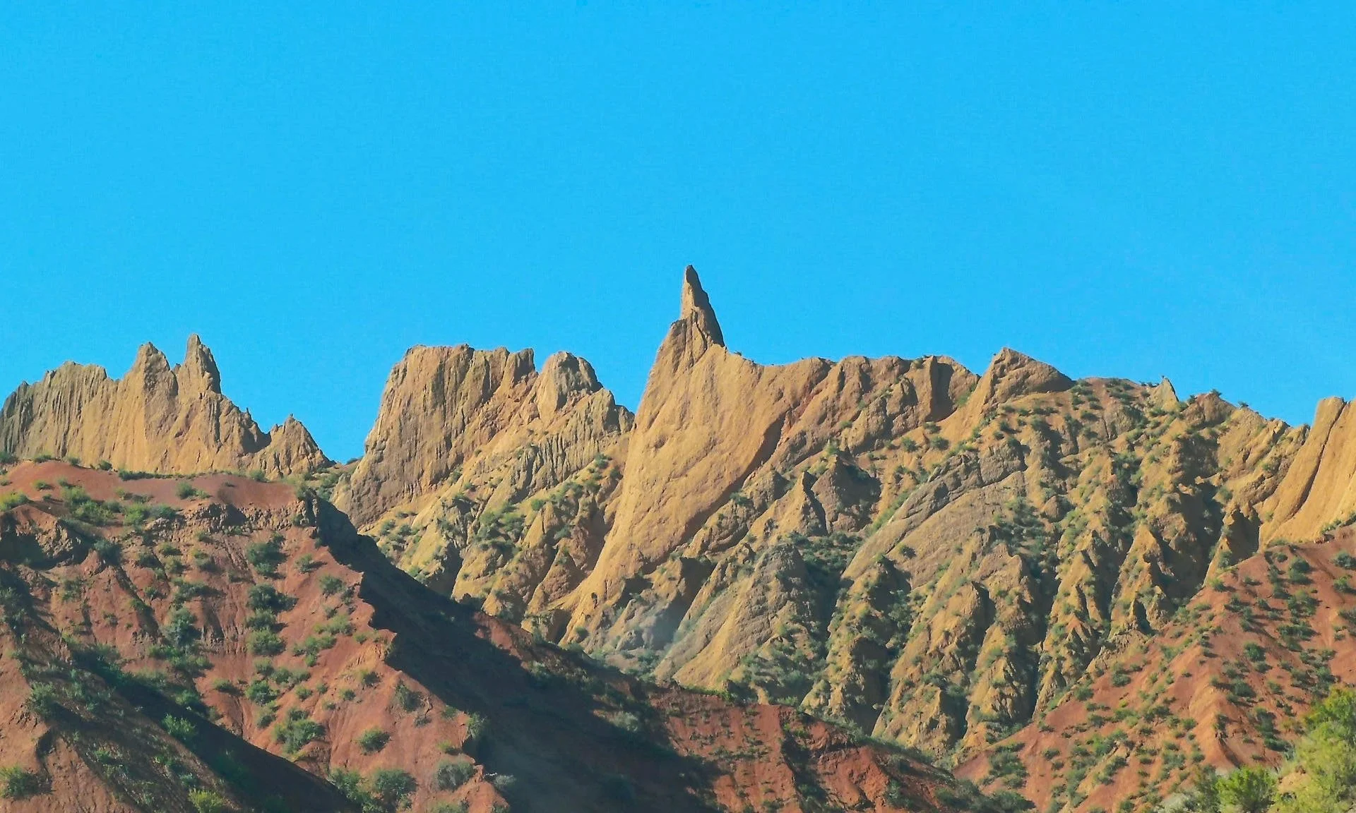 Colorful mountain range with sharp peaks, including one prominent pointed peak, under a clear blue sky.