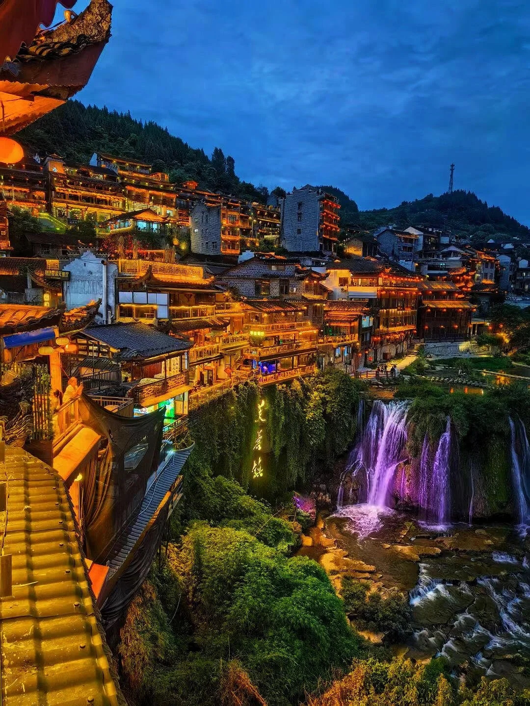Night view of a hillside town with buildings lit in warm lights, a waterfall illuminated with purple and pink lights, and lush greenery at the bottom.