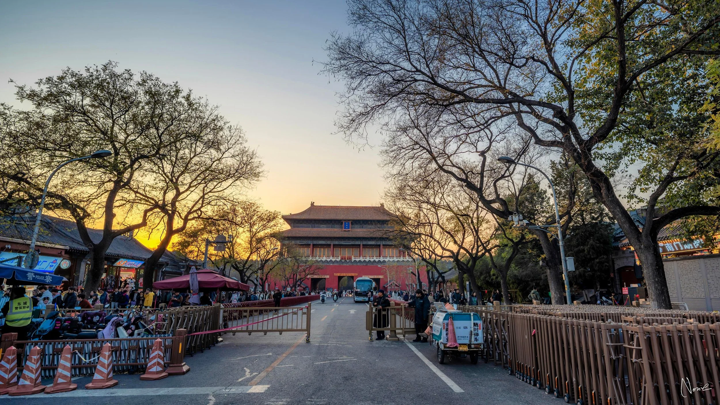 A busy street scene with people shopping and walking, traditional buildings, bare trees, and a historic gate during sunset.