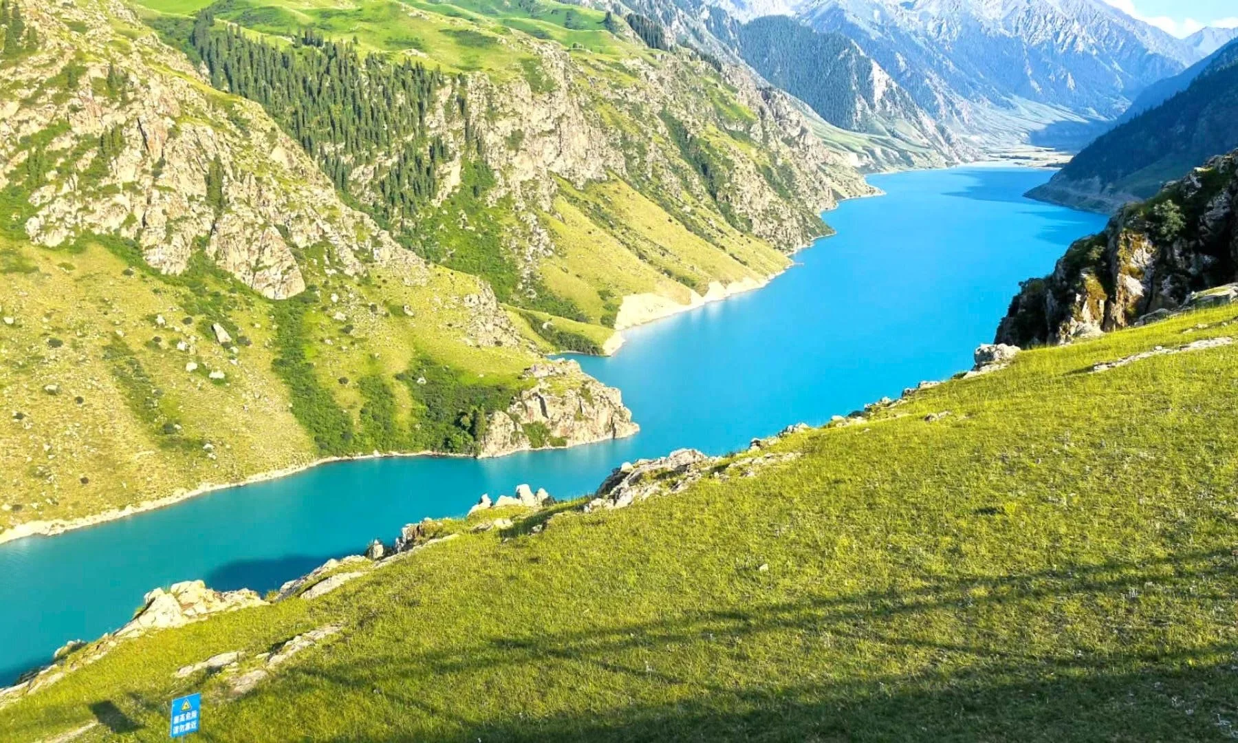 A scenic view of a large blue reservoir surrounded by lush green hills and steep mountains with rocky cliffs.