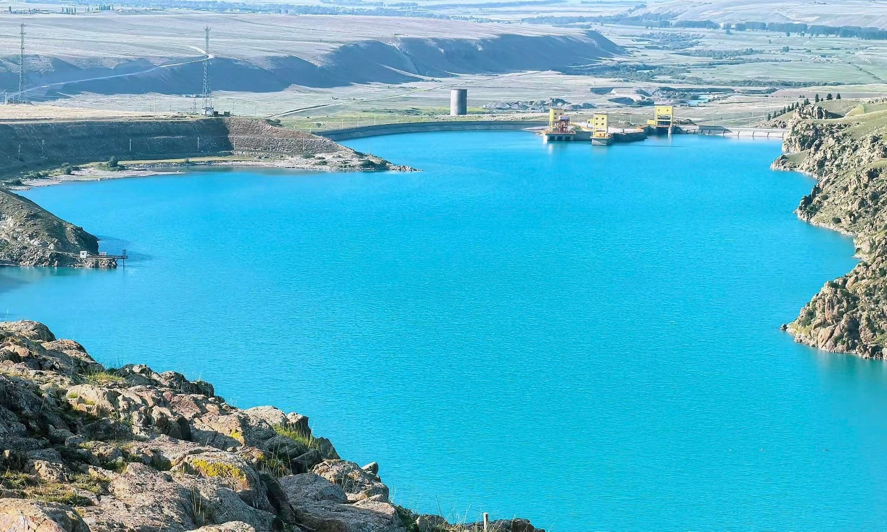 A large hydroelectric dam with a power station on a bright sunny day, with blue water in the reservoir and rocky terrain in the foreground.