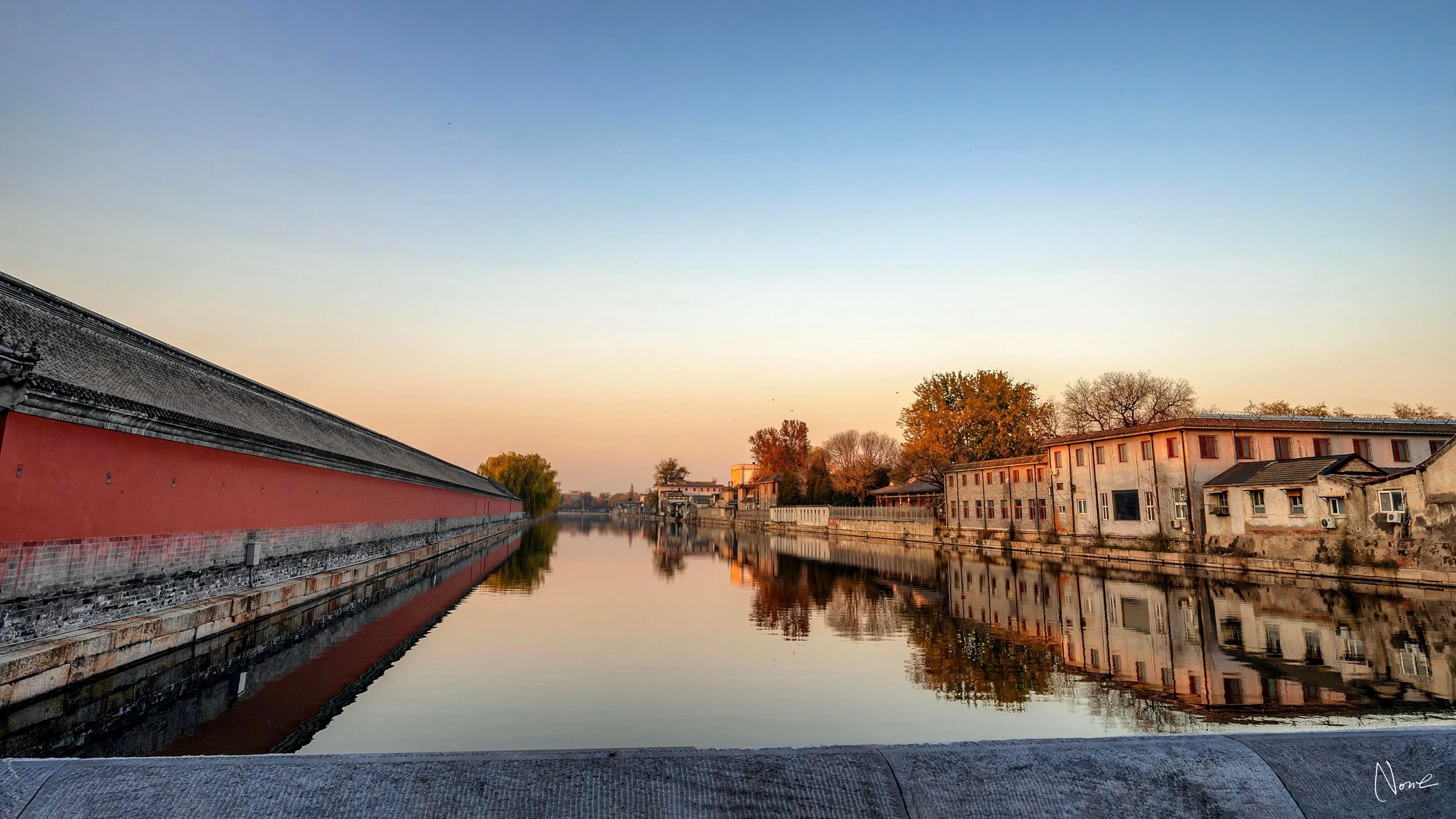 A peaceful canal scene at sunset with calm reflecting water, traditional Chinese architecture on the left, and modern buildings on the right, surrounded by trees with fall foliage.