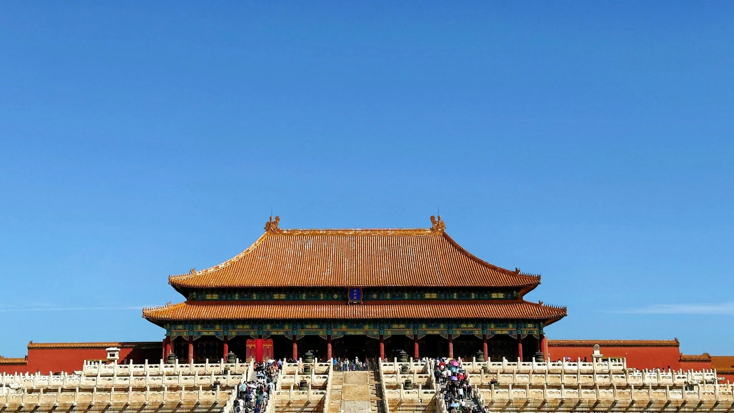 Traditional Chinese building with ornate roof, red pillars, and decorative roof ornaments, set against a clear blue sky, with a crowd of visitors in the courtyard.