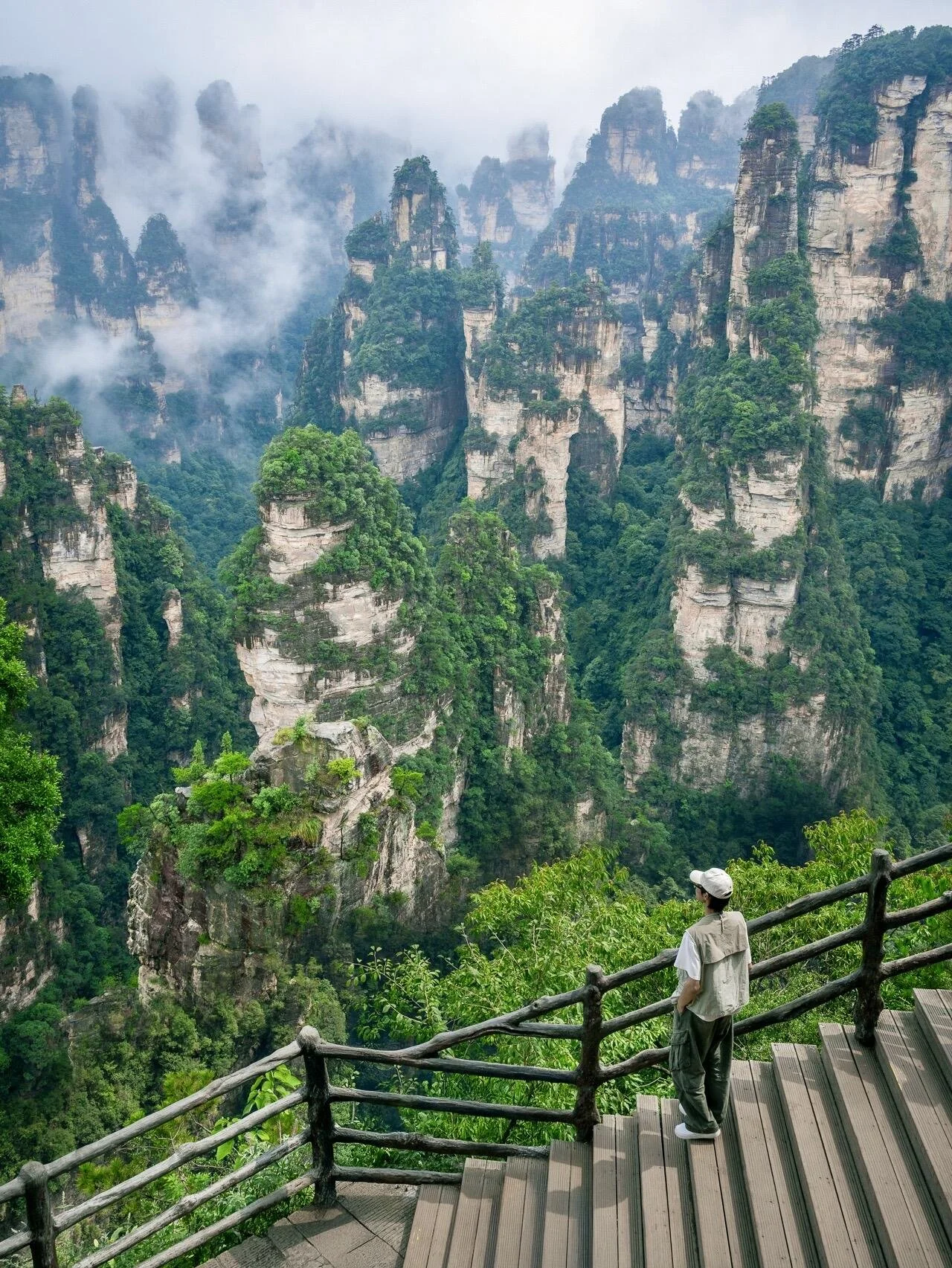 Person standing on a wooden viewing platform overlooking lush green mountains and towering rock formations shrouded in mist in Huangshan, China.